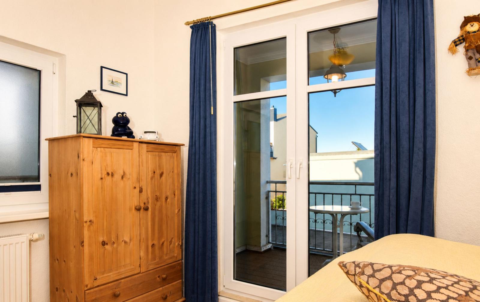 Zimmer mit Holzschrank, Balkon und blauen Vorhängen. Sicht auf Terrasse mit Tisch.