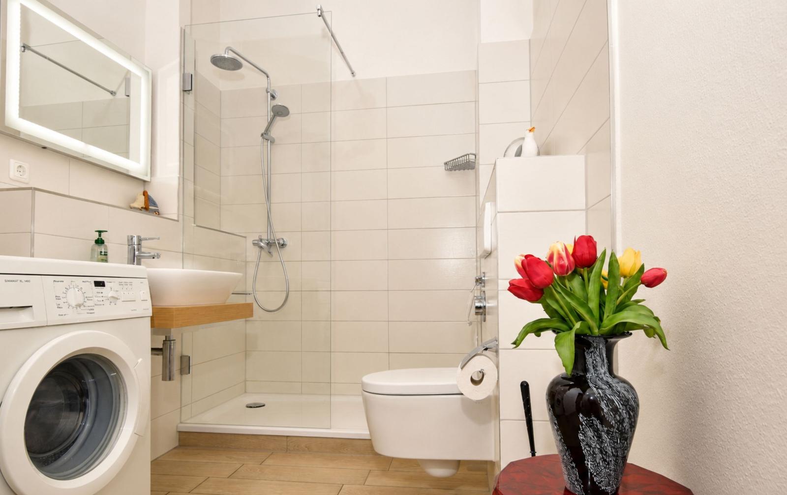 Badezimmer mit Dusche, Toilette, Waschmaschine und Blumenarrangement.