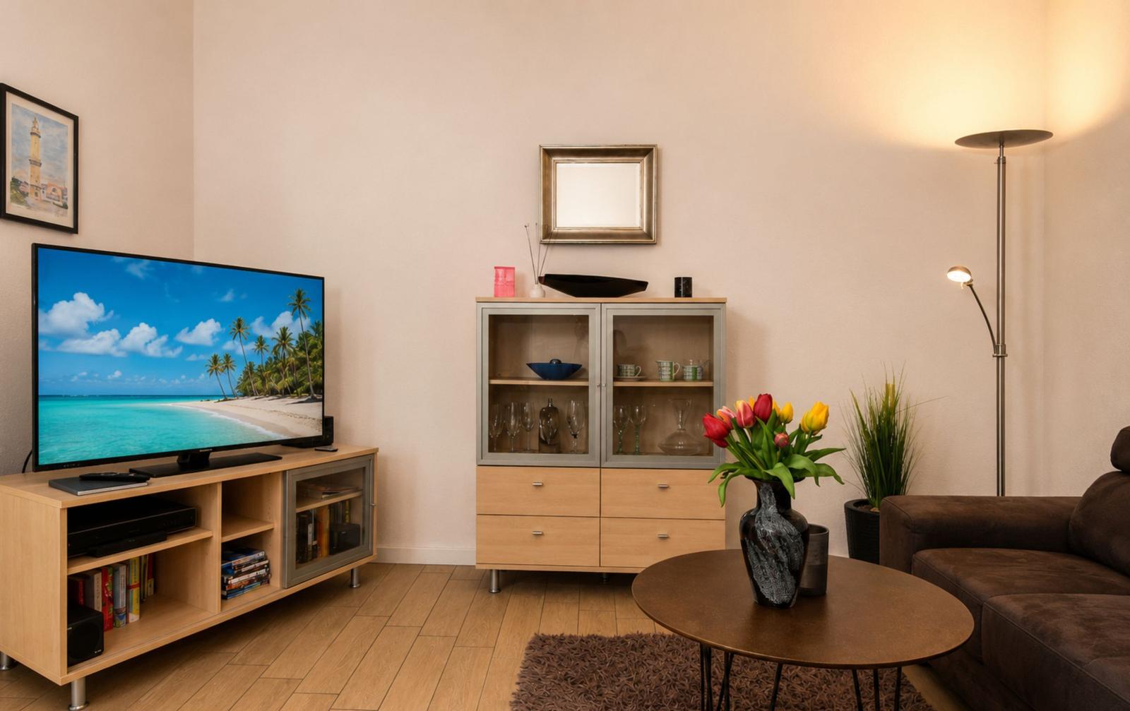 Wohnzimmer mit Fernseher, Couch und Glasvitrine. Auf dem Bildschirm zeigt sich eine Strandlandschaft.