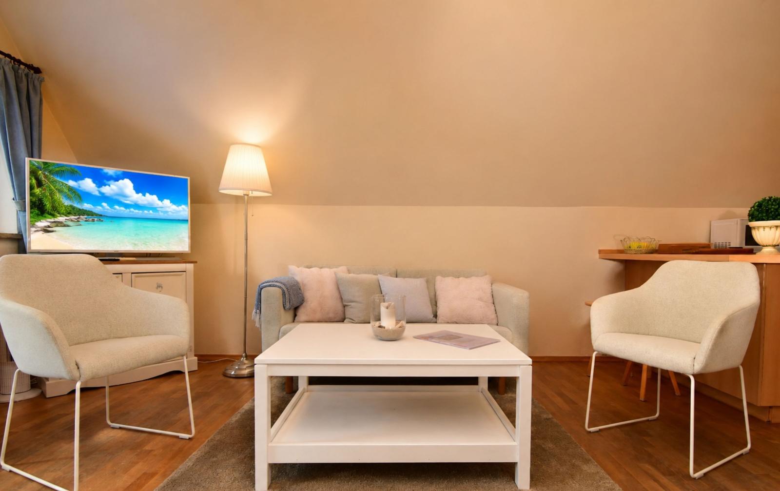 Wohnzimmer mit Sofa, zwei Sesseln, Tisch und Fernseher. Holzboden und Wandfarbe beige.