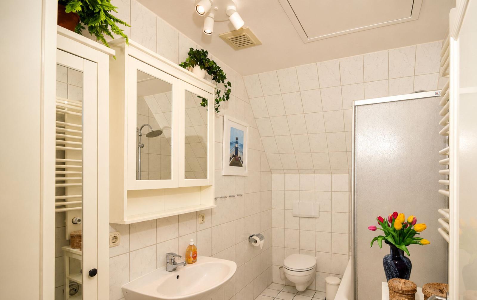 Badezimmer mit Dusche, Toilette und Waschbecken. Weiße Fliesen und Pflanzen.