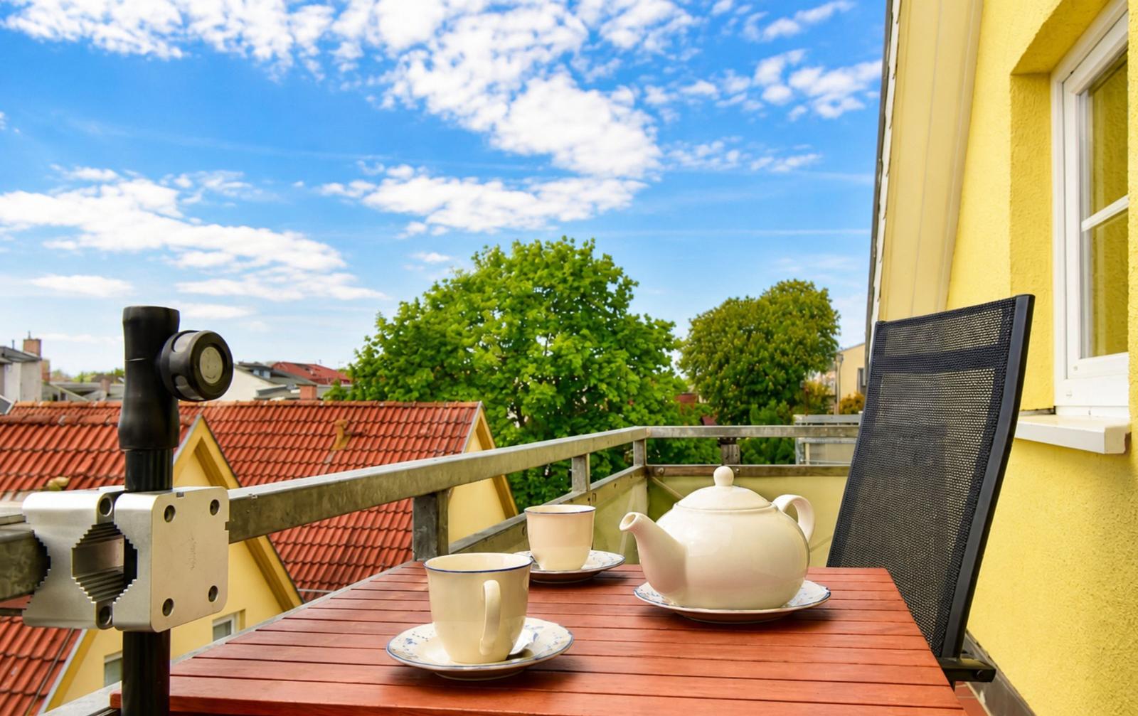 Terrasse mit Tisch, Teekanne und zwei Tassen. Blick auf Dächer und Bäume unter blauem Himmel.