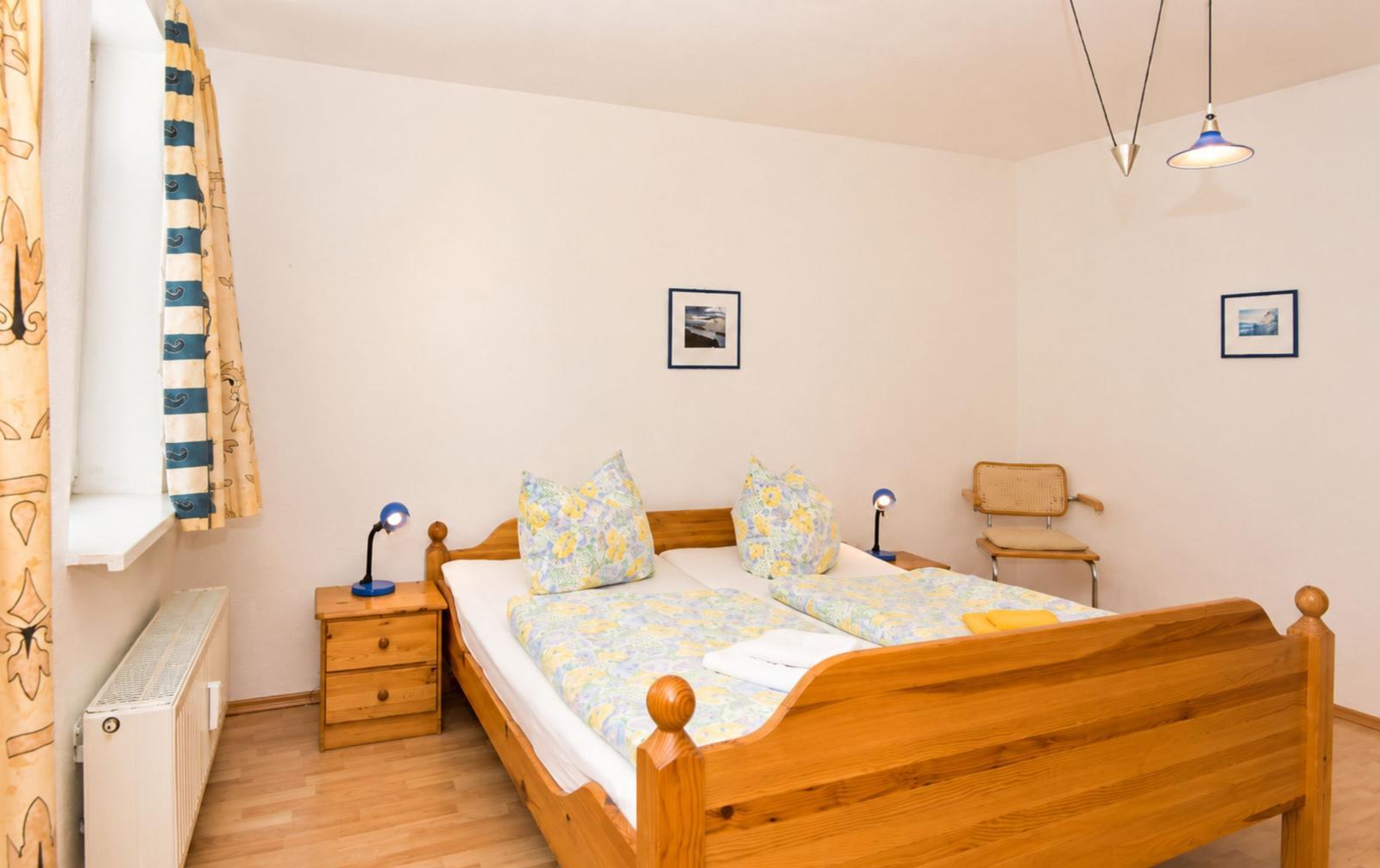 Ein Schlafzimmer mit Holzbett, Nachttisch, Stuhl und Fenster mit Vorhängen.