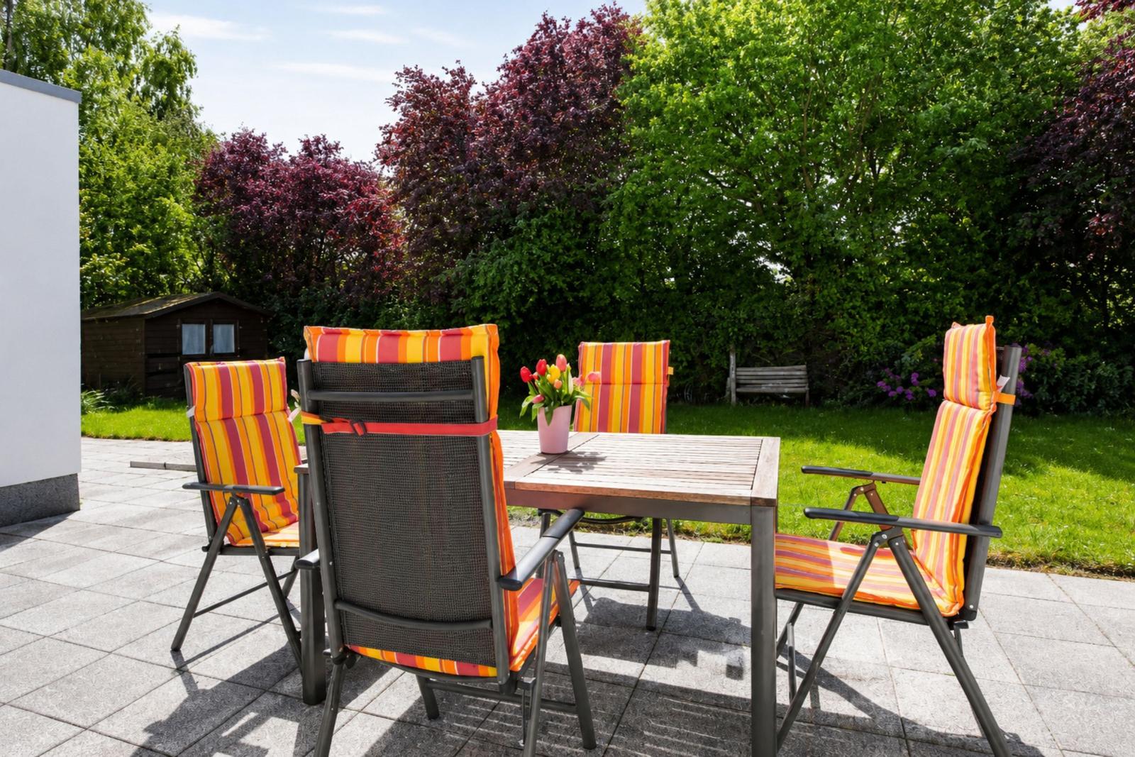 Terrasse mit Tisch und vier Stühlen mit orangefarbenen Kissen. Hintergrund: grüner Garten und Bäume.