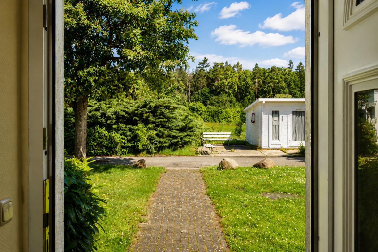 Durch die Tür blickt man auf einen gepflasterten Weg, einen weißen Gartenhäuschen und einen Wald.