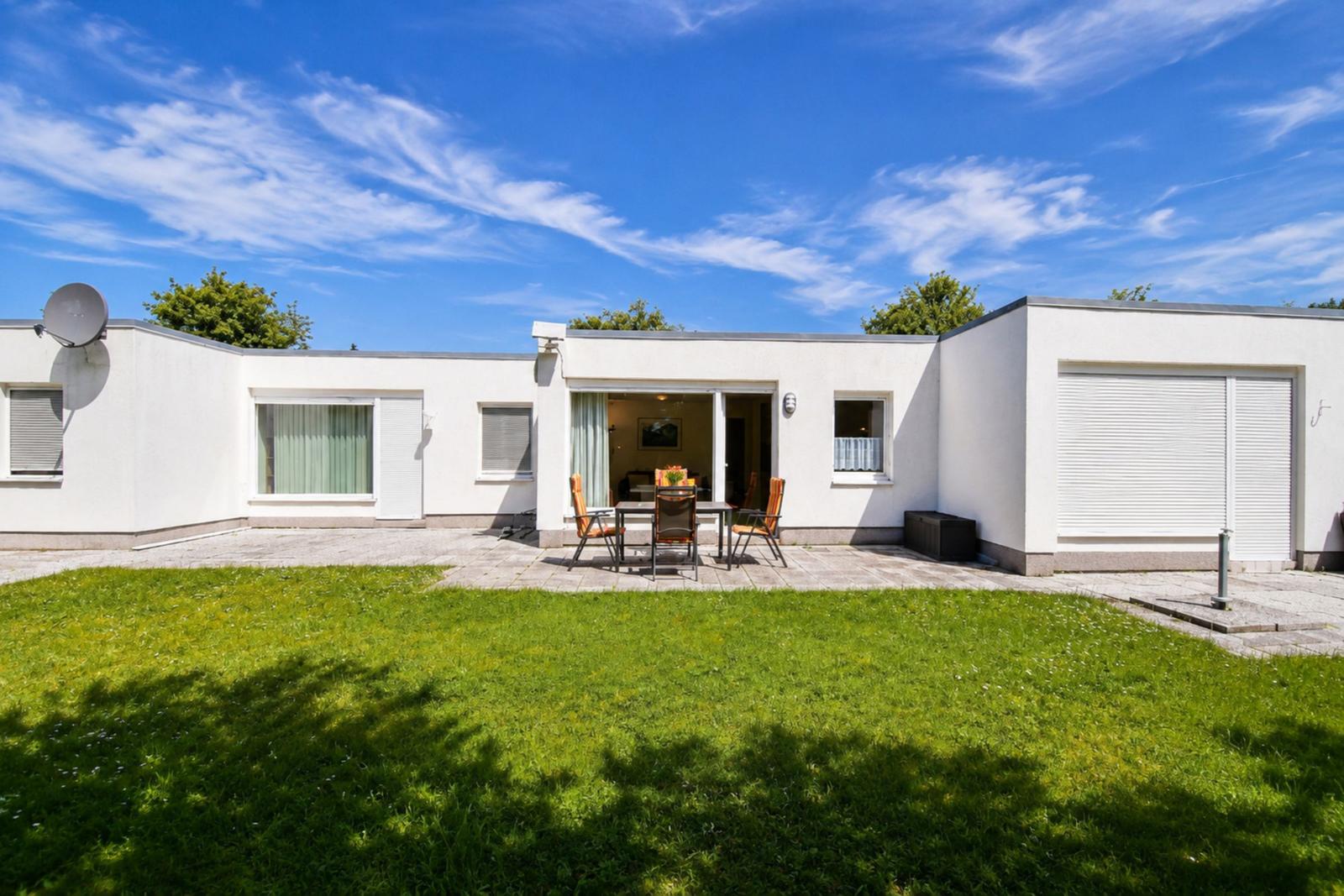 Modernes weißes Haus mit Garten, Terrasse und Grillplatz unter blauem Himmel.
