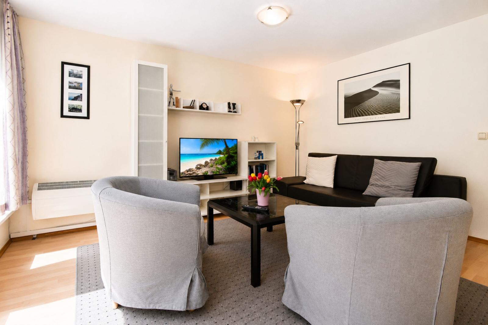 Wohnzimmer mit Sofa, zwei Sesseln, Fernseher und Tisch mit Blumen.