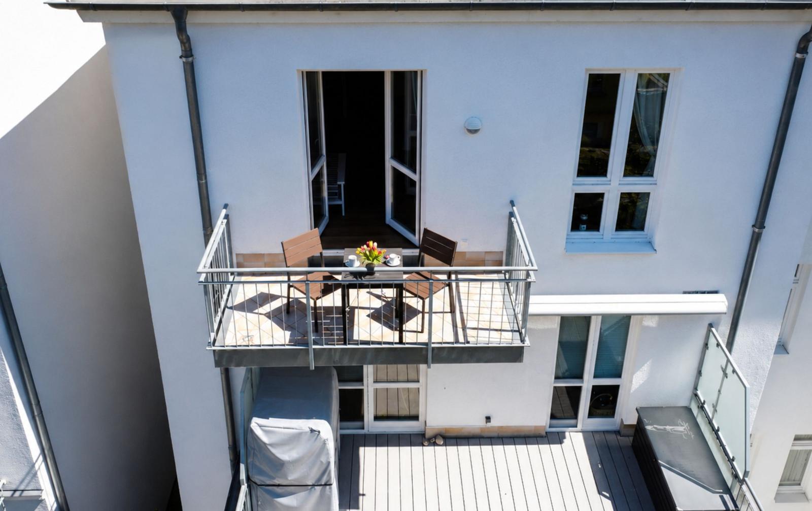 Terrace with table, two chairs, and flower arrangement. Background: living area with windows.