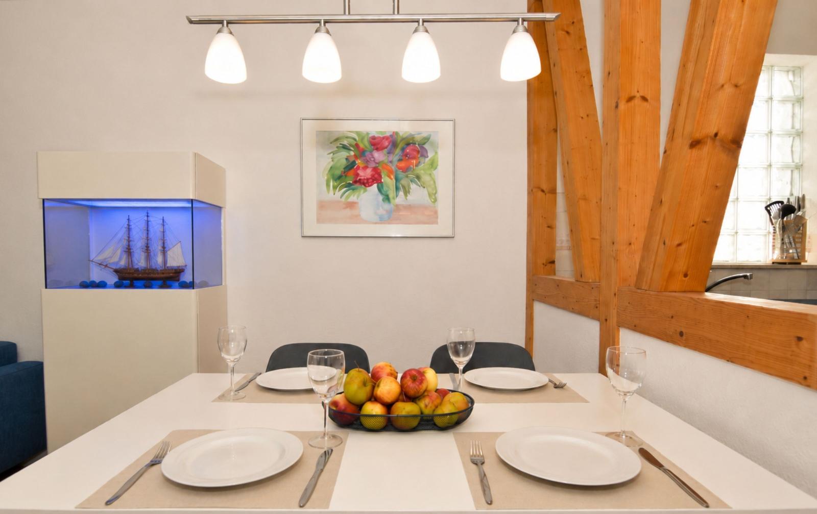 Dining room with table, chairs, and fruit bowl. Background: wall art and wooden beams.