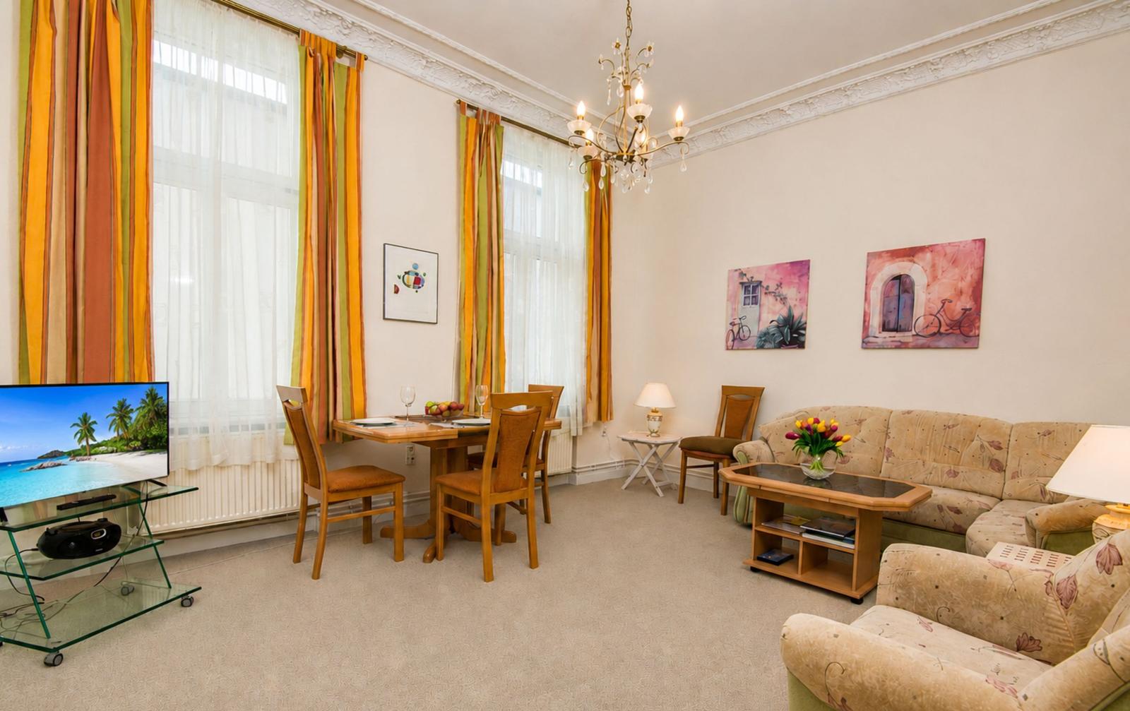 Wohn- und Essbereich mit Sofa, Tisch, Stühlen und TV. Fenster mit bunten Vorhängen.