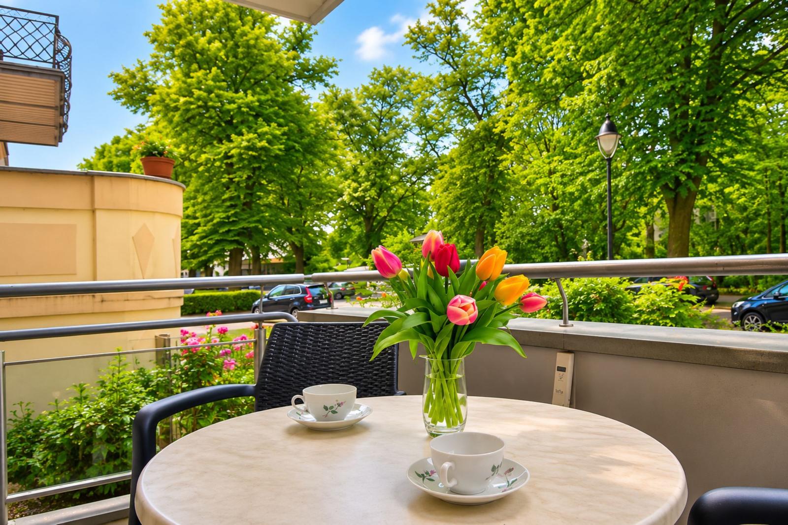 Balcony with table, chairs, and tulips. View of green trees and street.