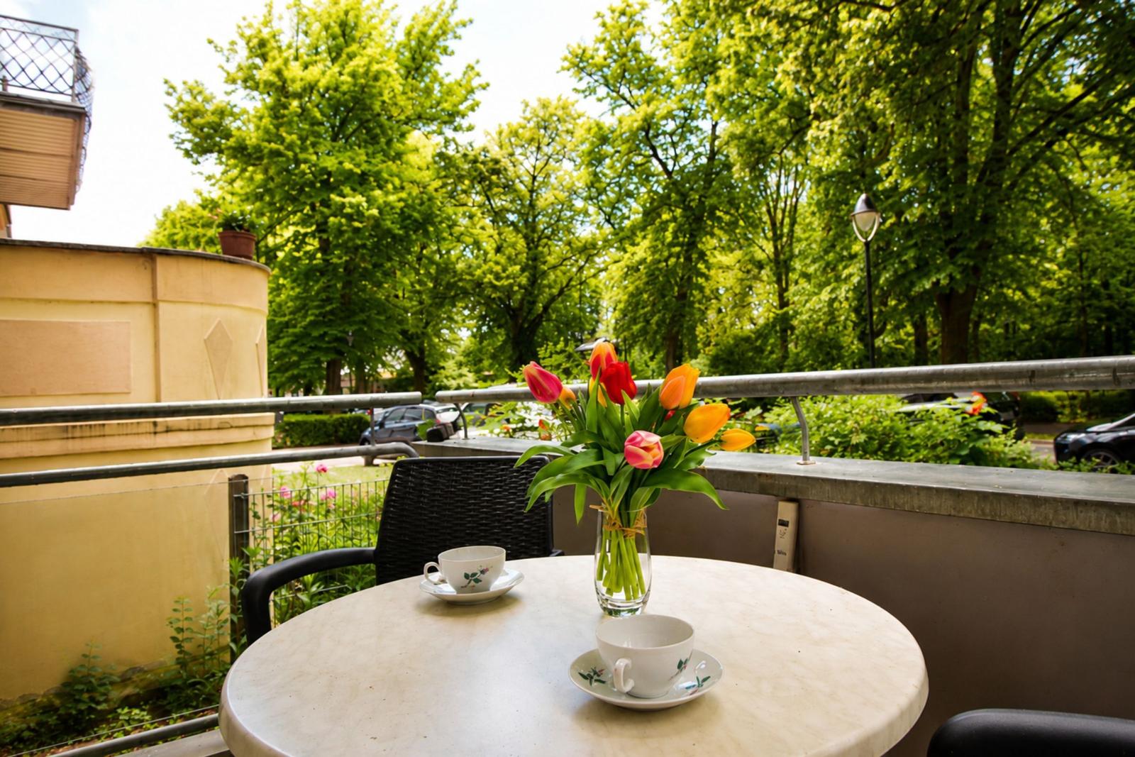 Round table with two cups and flowers on balcony with view of green trees.