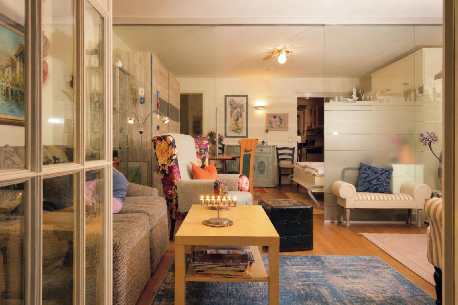 Living room with sofa, table, and chairs. Glass wall separates space. Rug and cushions visible.