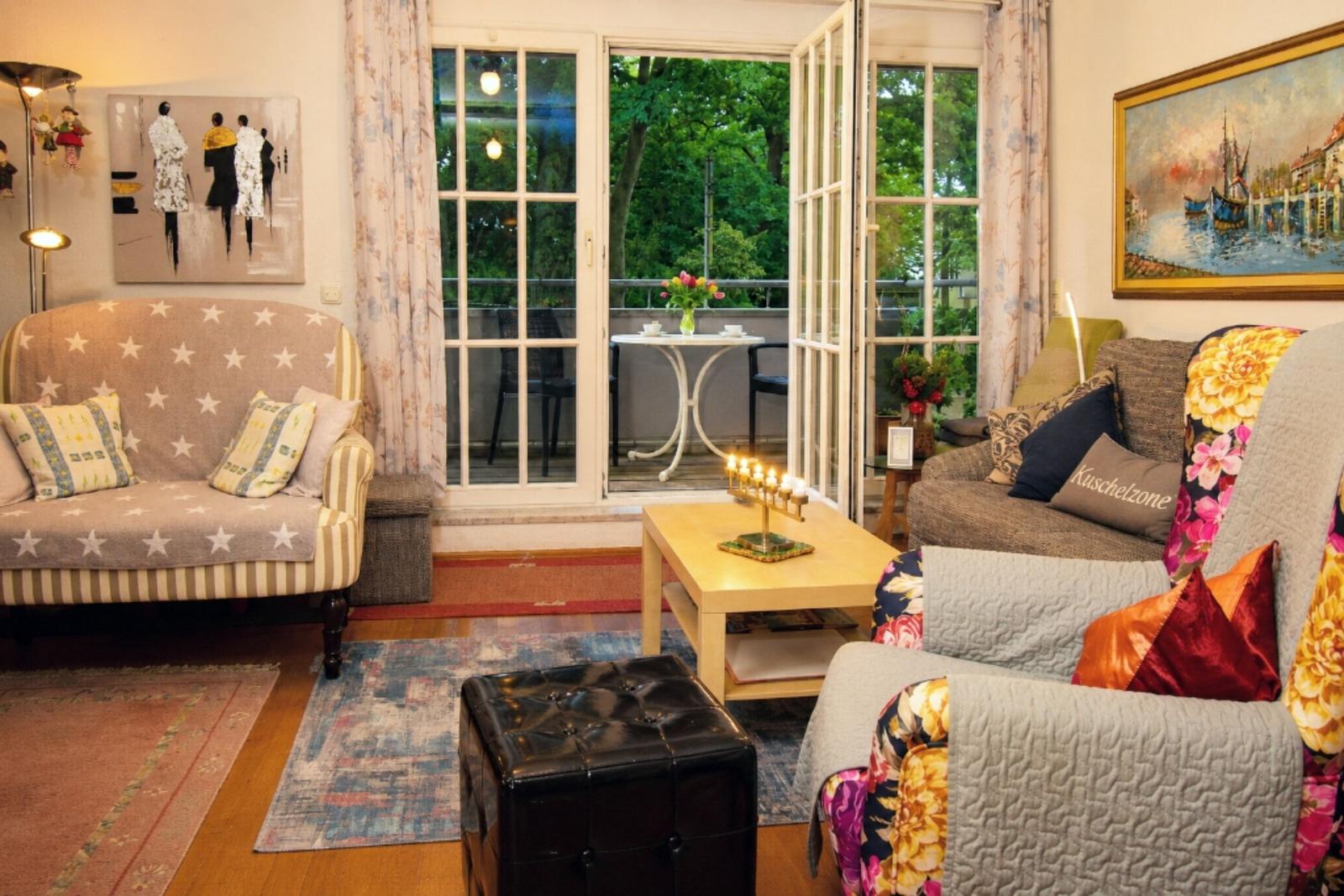 Living room with sofa, armchair, and balcony. Windows open, view of greenery.