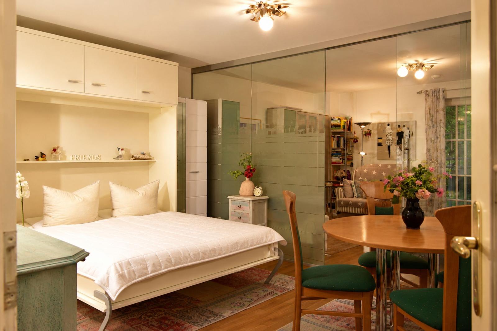 Bedroom with bed, wardrobe, and seating area. Glass partition separates space. Flowers and furniture visible.