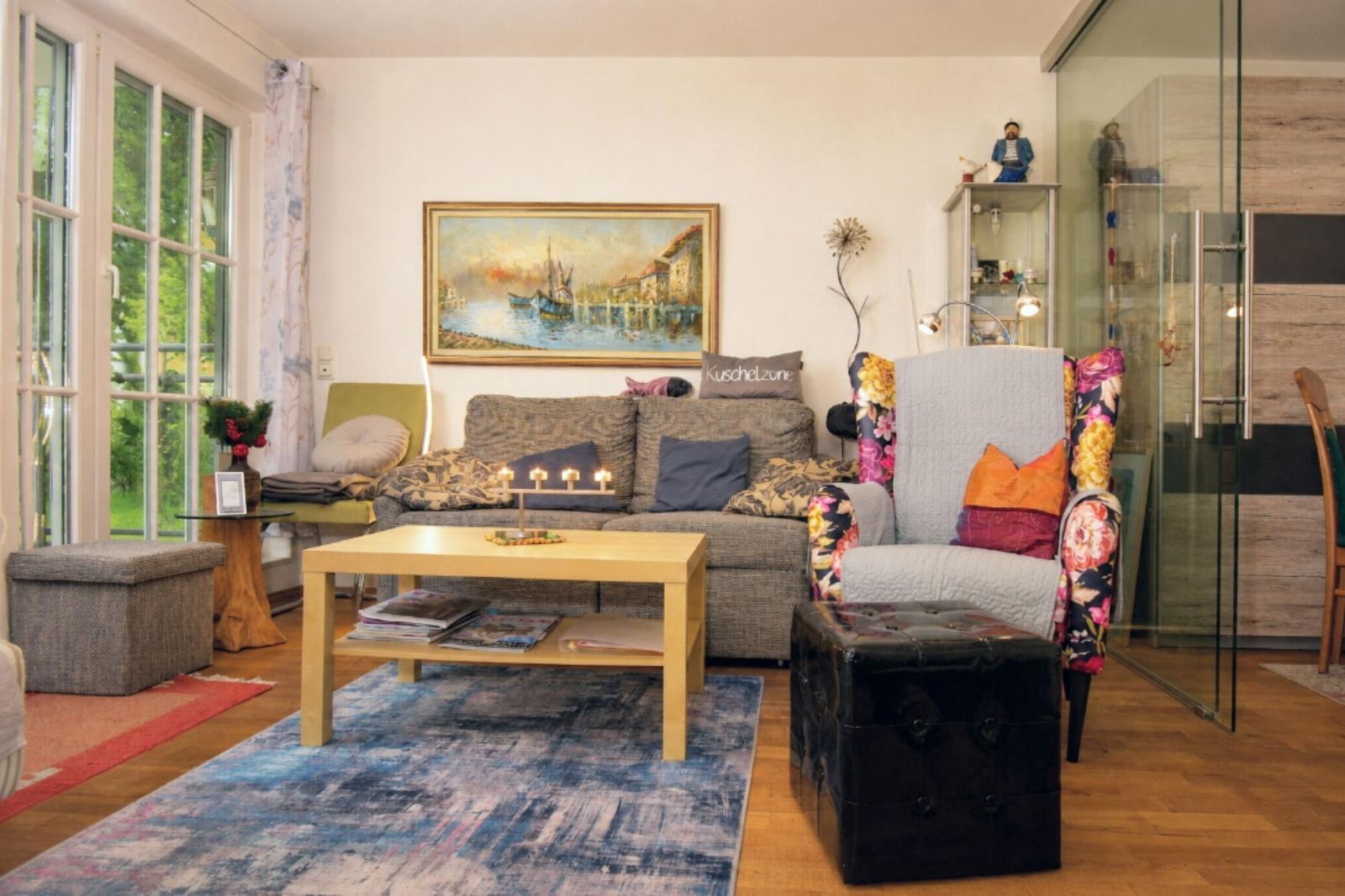 Living room with sofa, armchair, and table. Windows, wall painting, and rug are visible.