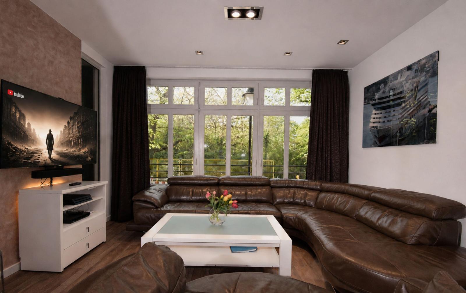 Ein Wohnzimmer mit braunem Ledersofa, Glas-Tisch und TV an der Wand.