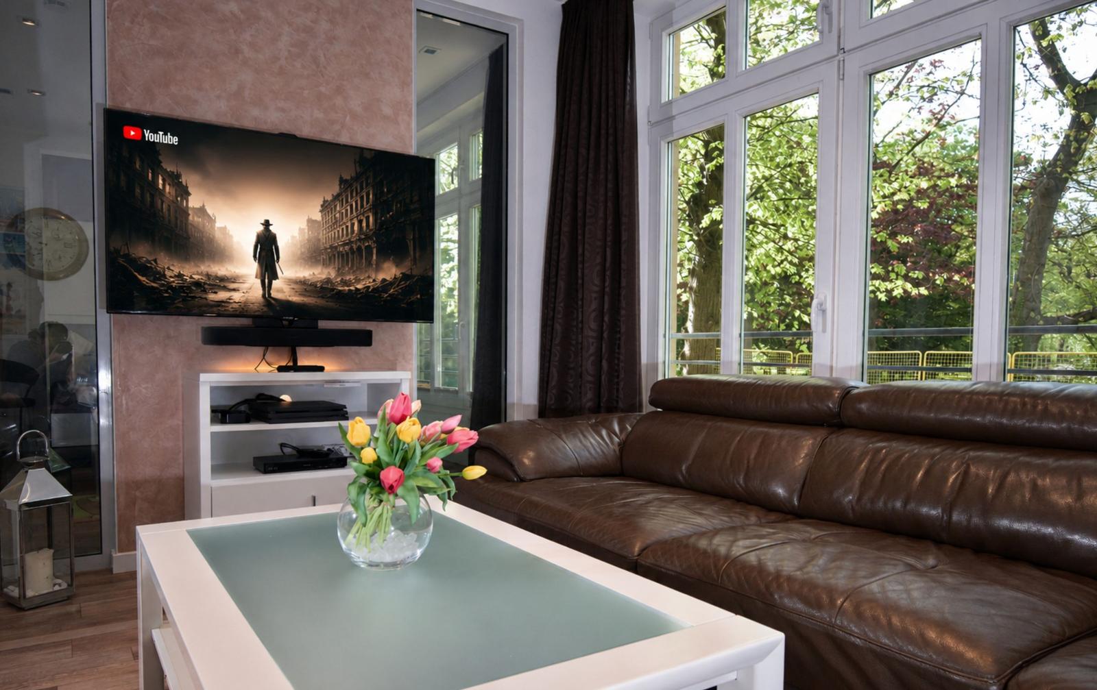 Ein Wohnzimmer mit braunem Ledersofa, Fernseher und großem Fenster mit Blick auf Bäume.