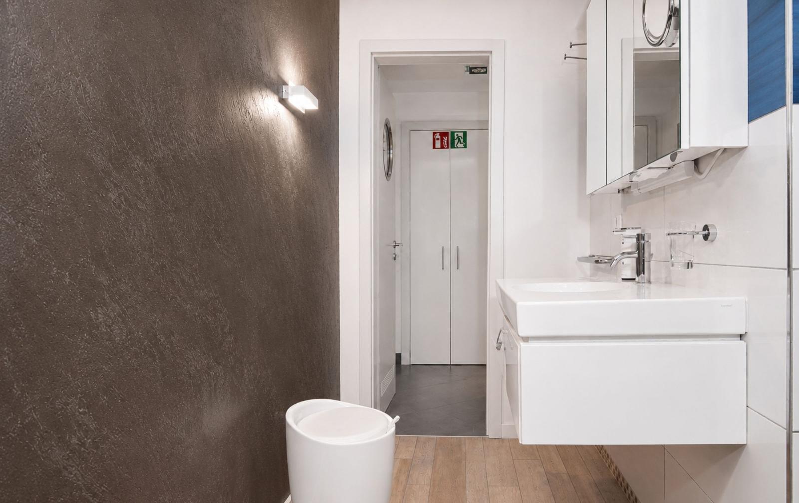 Badezimmer mit Waschbecken, Spiegel und Toilette. Holzfußboden und dunkle Wand.