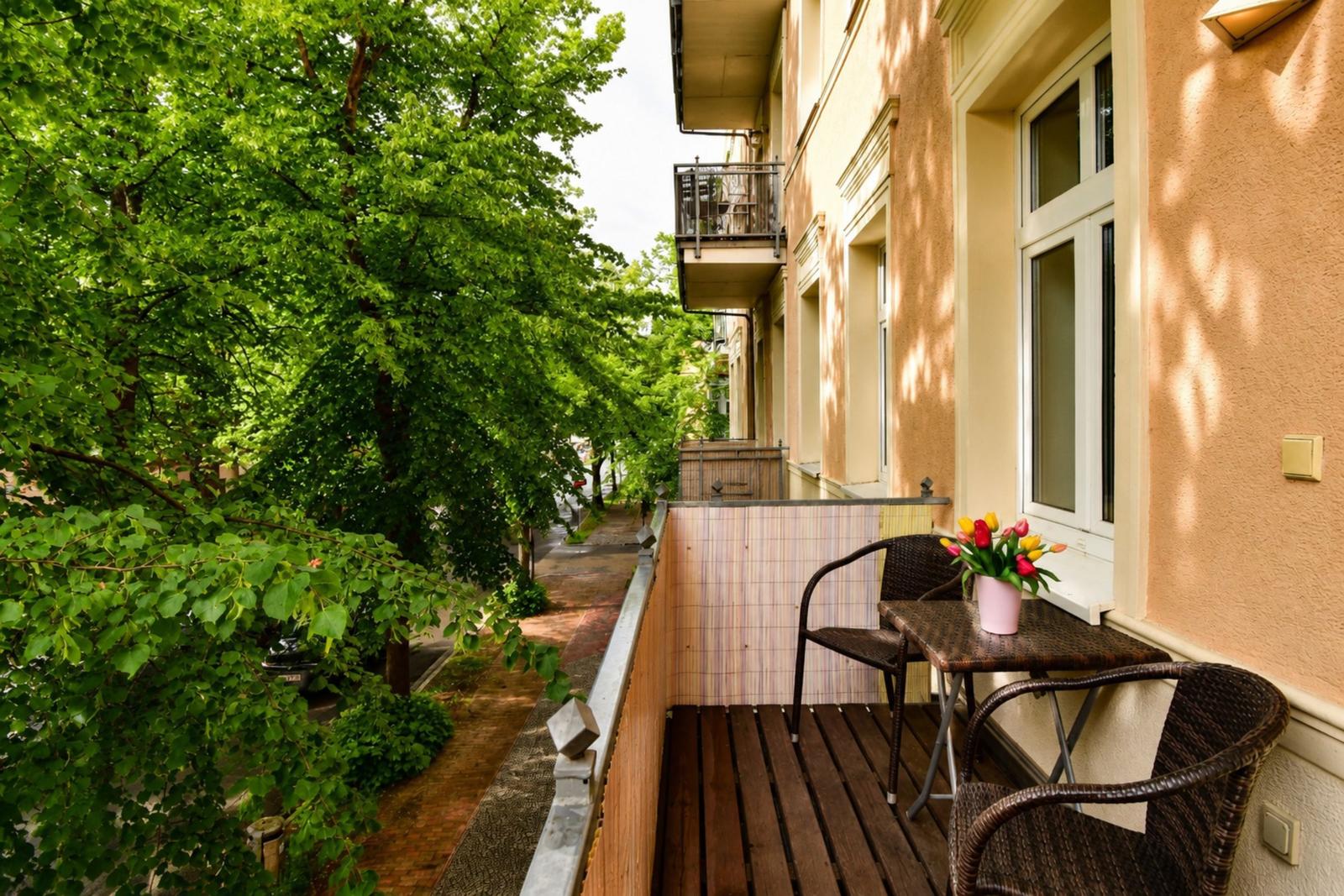 Terrasse mit Tisch, zwei Stühlen und Blumenstrauß. Blick auf grüne Bäume und Gehweg.