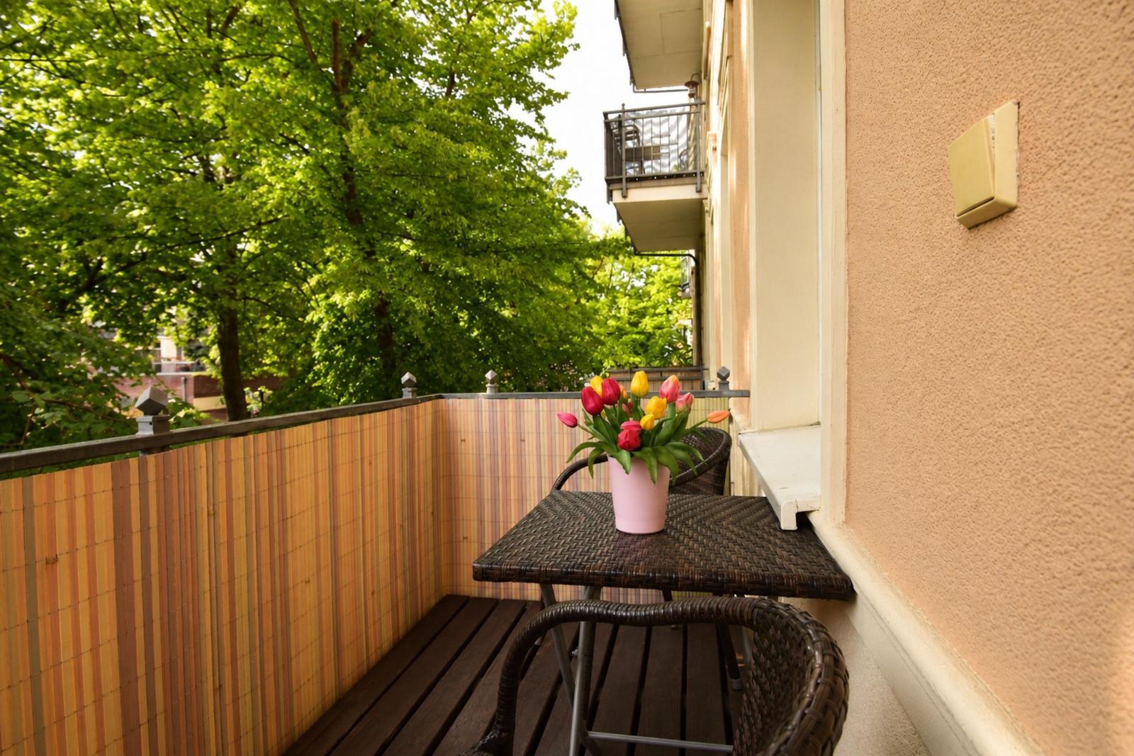 Balcony with table, chairs, and tulips. Wooden floor and bamboo wall. Green trees in background.