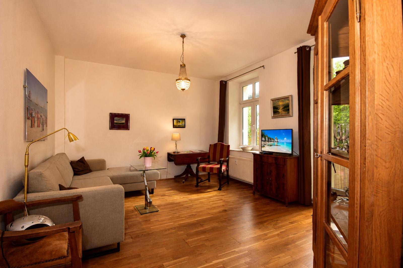 Wohnzimmer mit Sofa, Fernseher und Holzboden. Fenster mit Blick auf Grün.