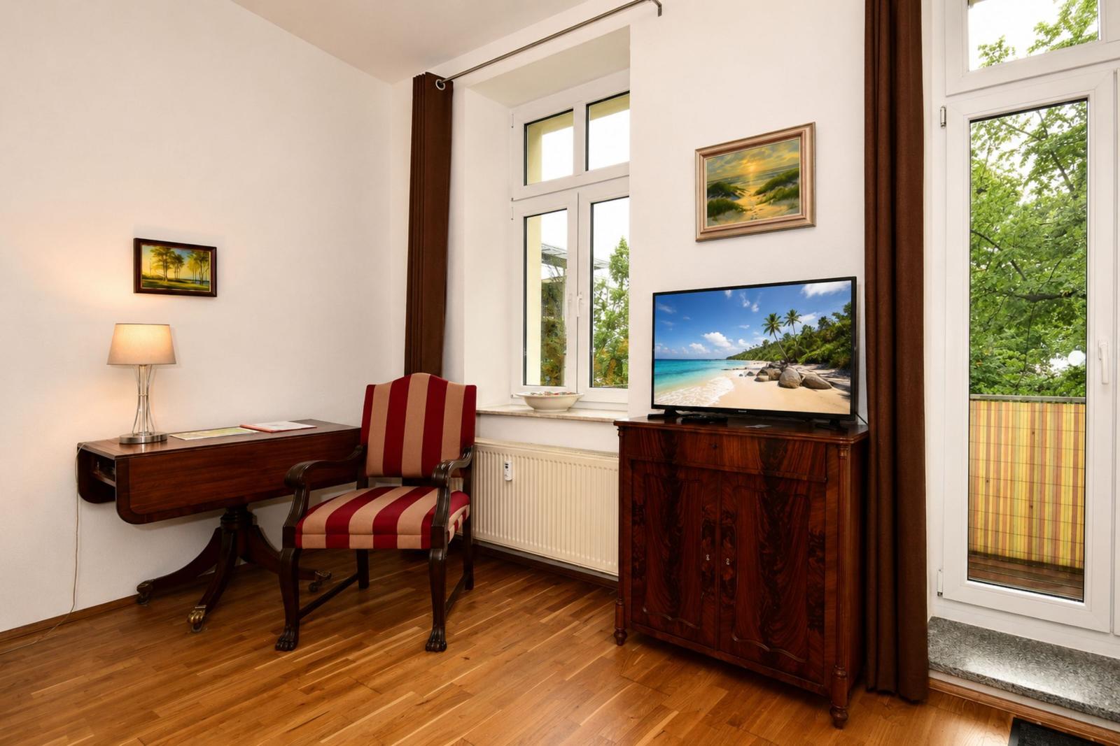 Ein Raum mit Schreibtisch, Stuhl und Fernseher auf einem Sideboard. Fenster mit Blick auf Grün.