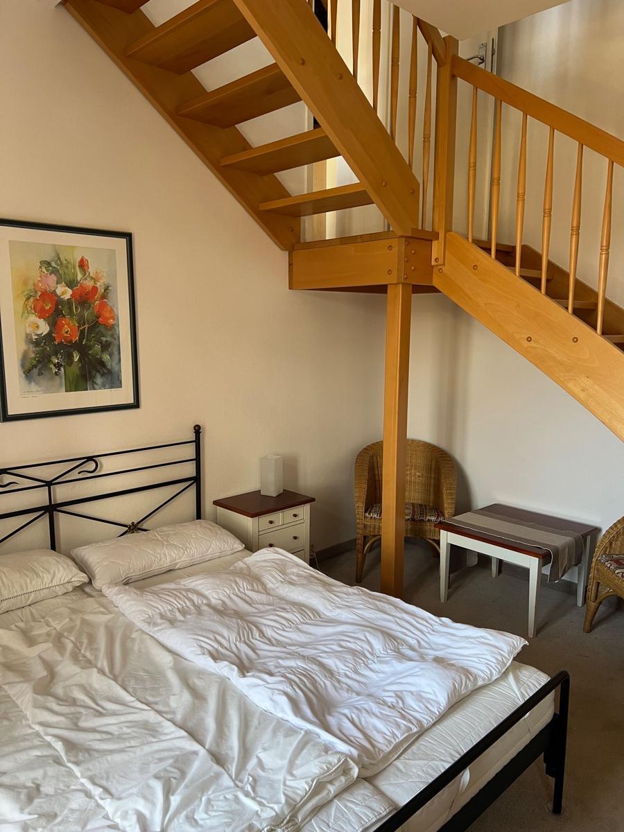 Schlafzimmer mit Doppelbett, Holztreppe und Wickerstuhl.