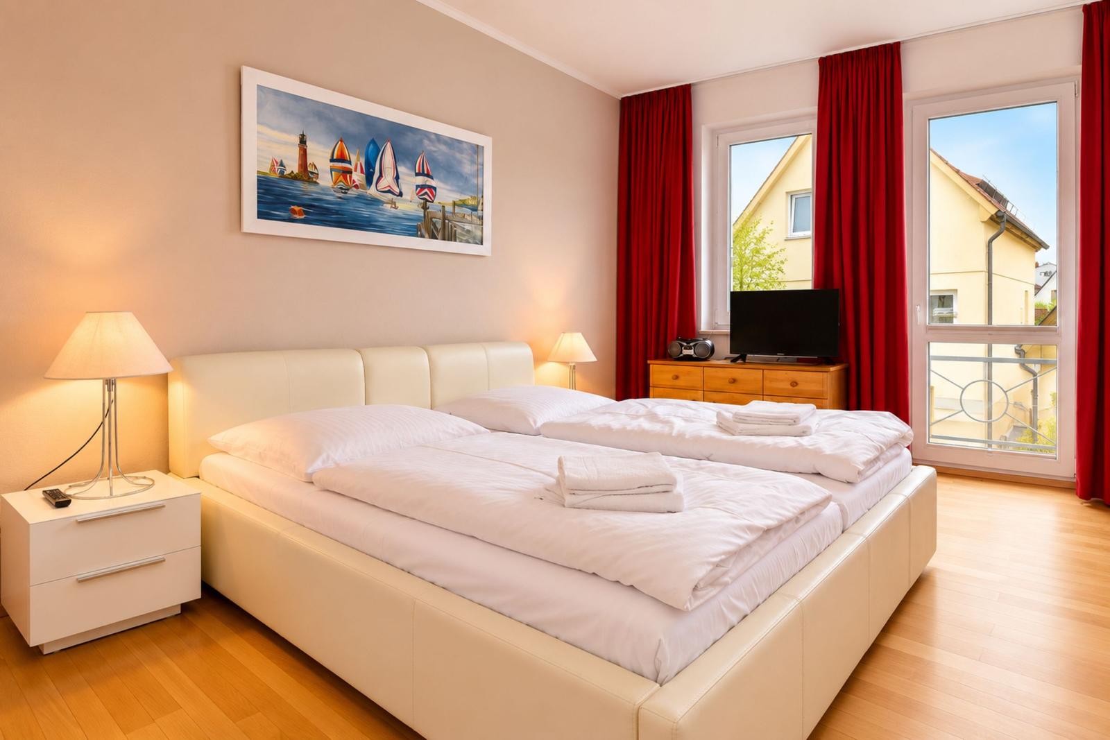 Schlafzimmer mit Doppelbett, Fenster und roten Vorhängen. Wandbild und Nachttische.
