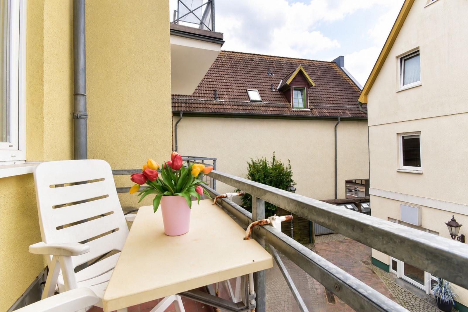 Balcony with white chair, table, and pink pot with tulips. View of neighboring buildings.