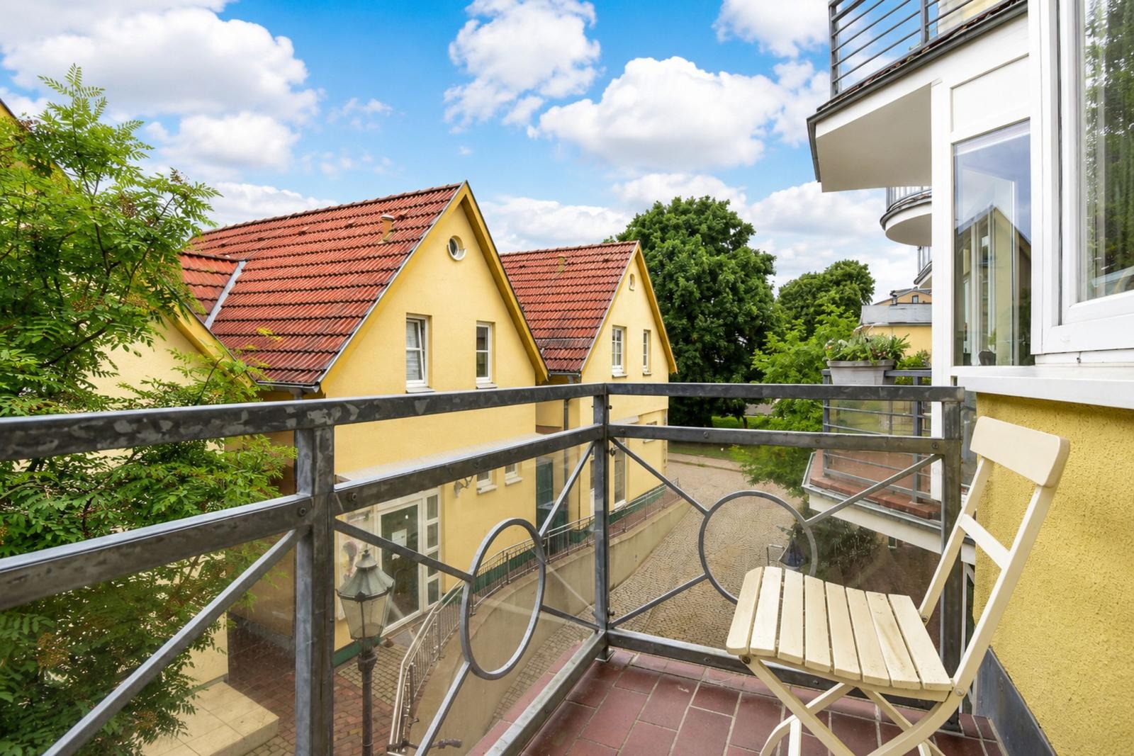 Balkon mit Stuhl, Blick auf gelbe Häuser und Bäume unter blauem Himmel.