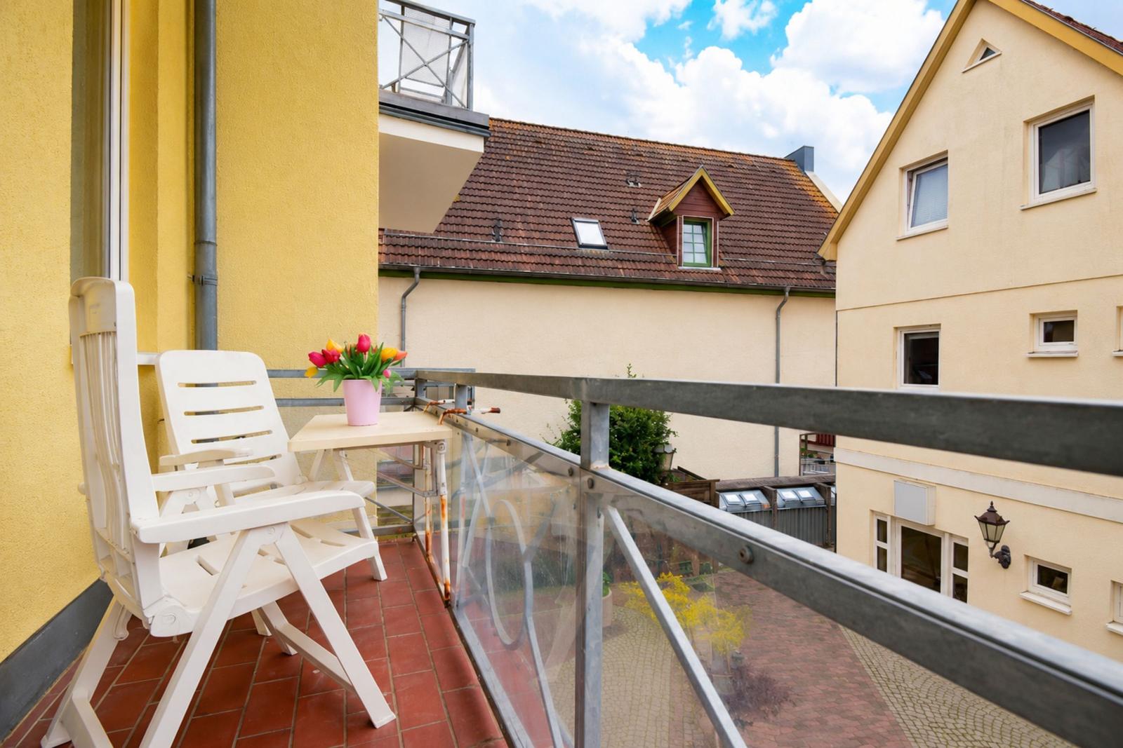 Balkon mit weißen Stühlen und Tisch, Blumen in Vase, Blick auf Dächer und Hauswand.