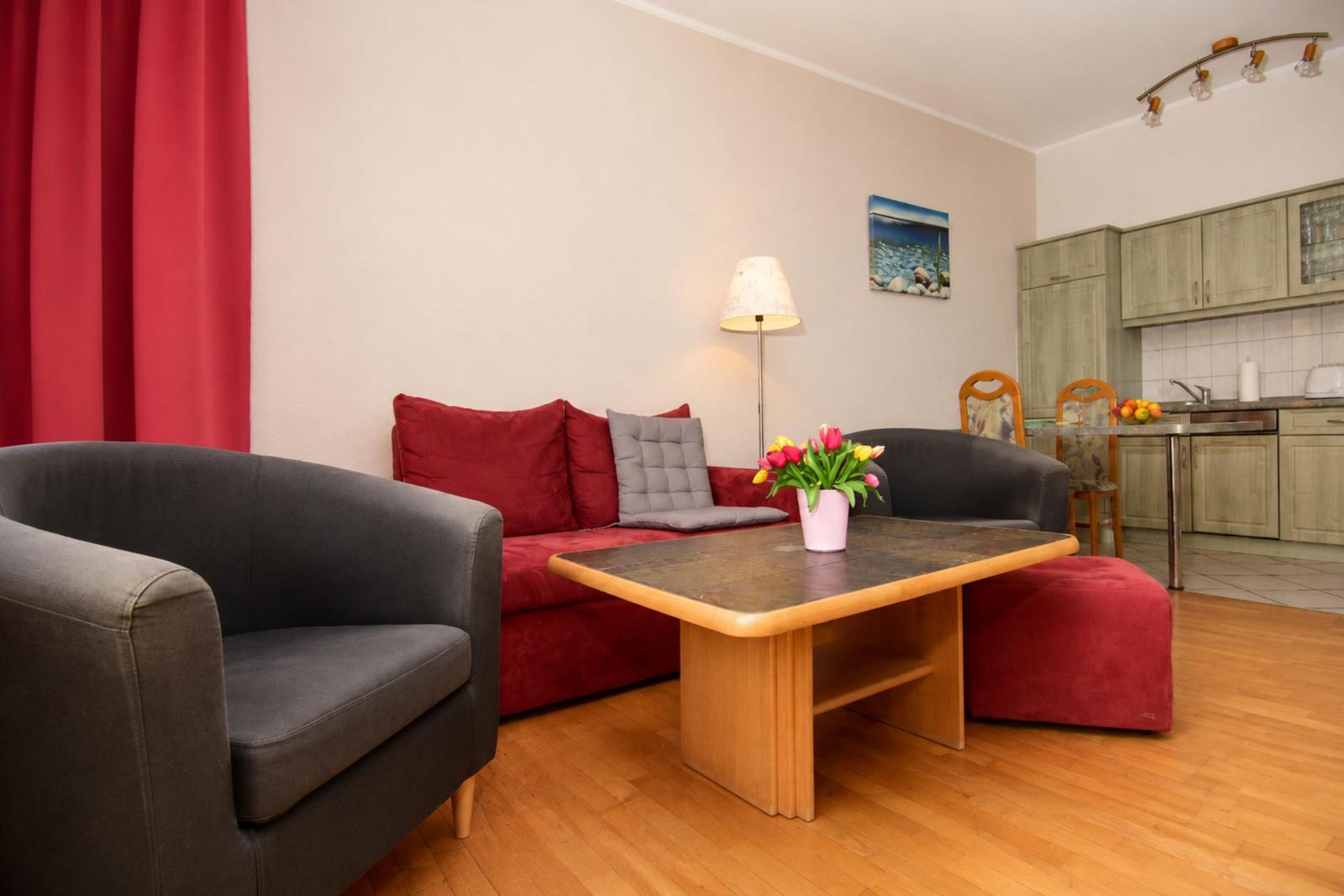 Wohnbereich mit rotem Sofa, grauen Sesseln und Holztisch. Küche im Hintergrund.
