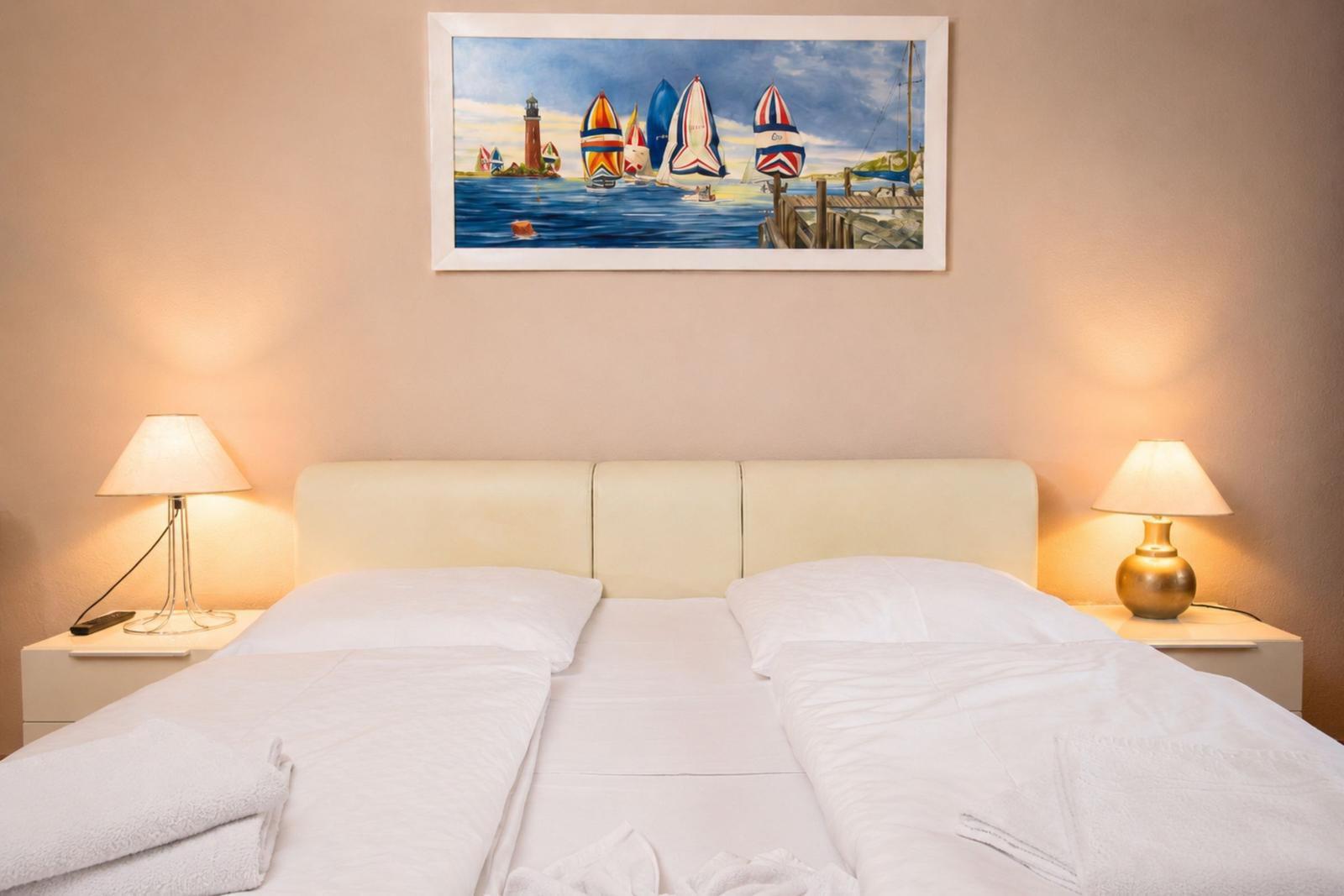 Doppelbett mit weißen Laken, zwei Nachttische mit Lampen und Wandbild mit Segelbooten.
