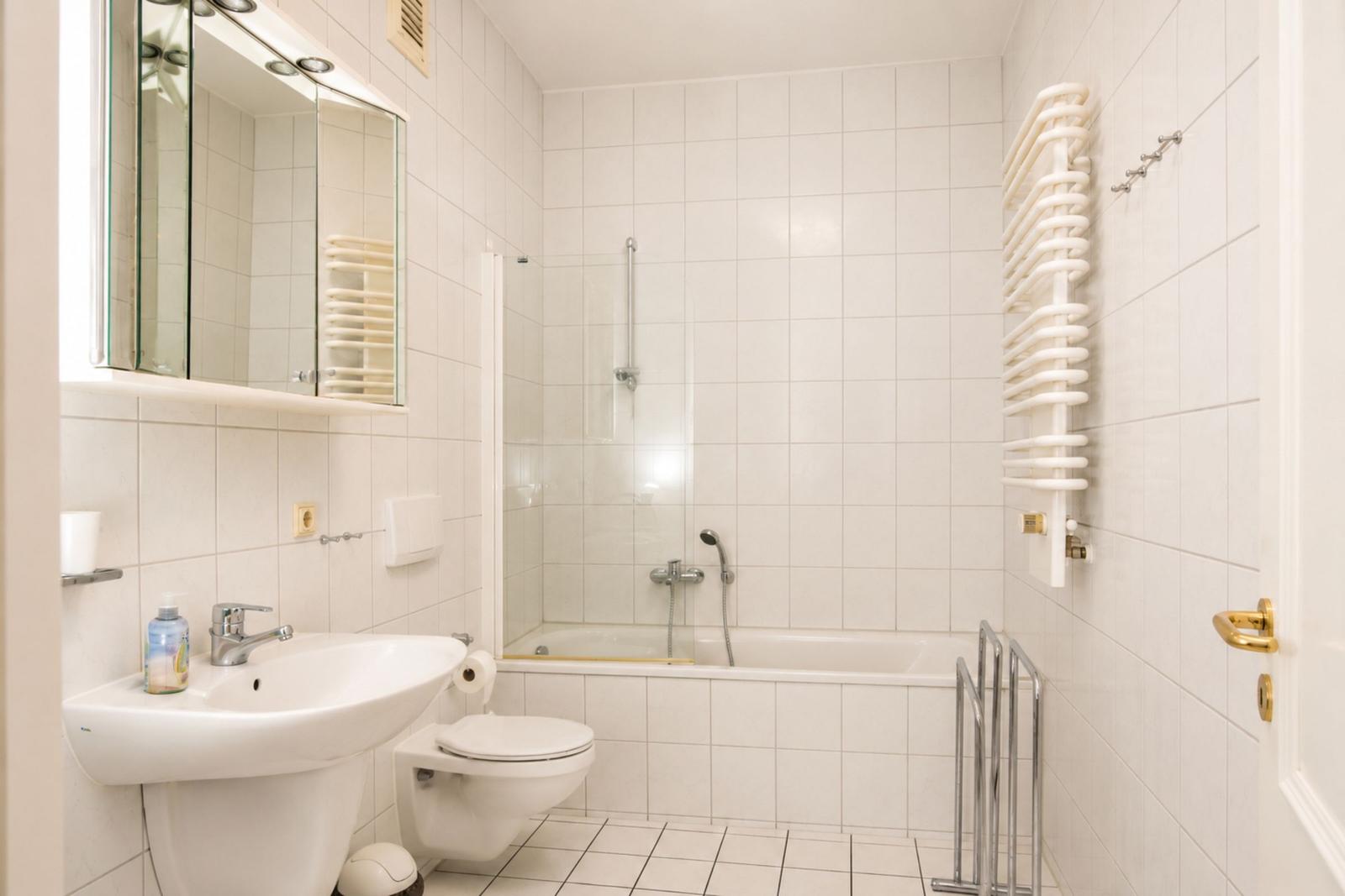 Badezimmer mit Dusche, Waschbecken und Toilette. Weiße Fliesen und Heizkörper.