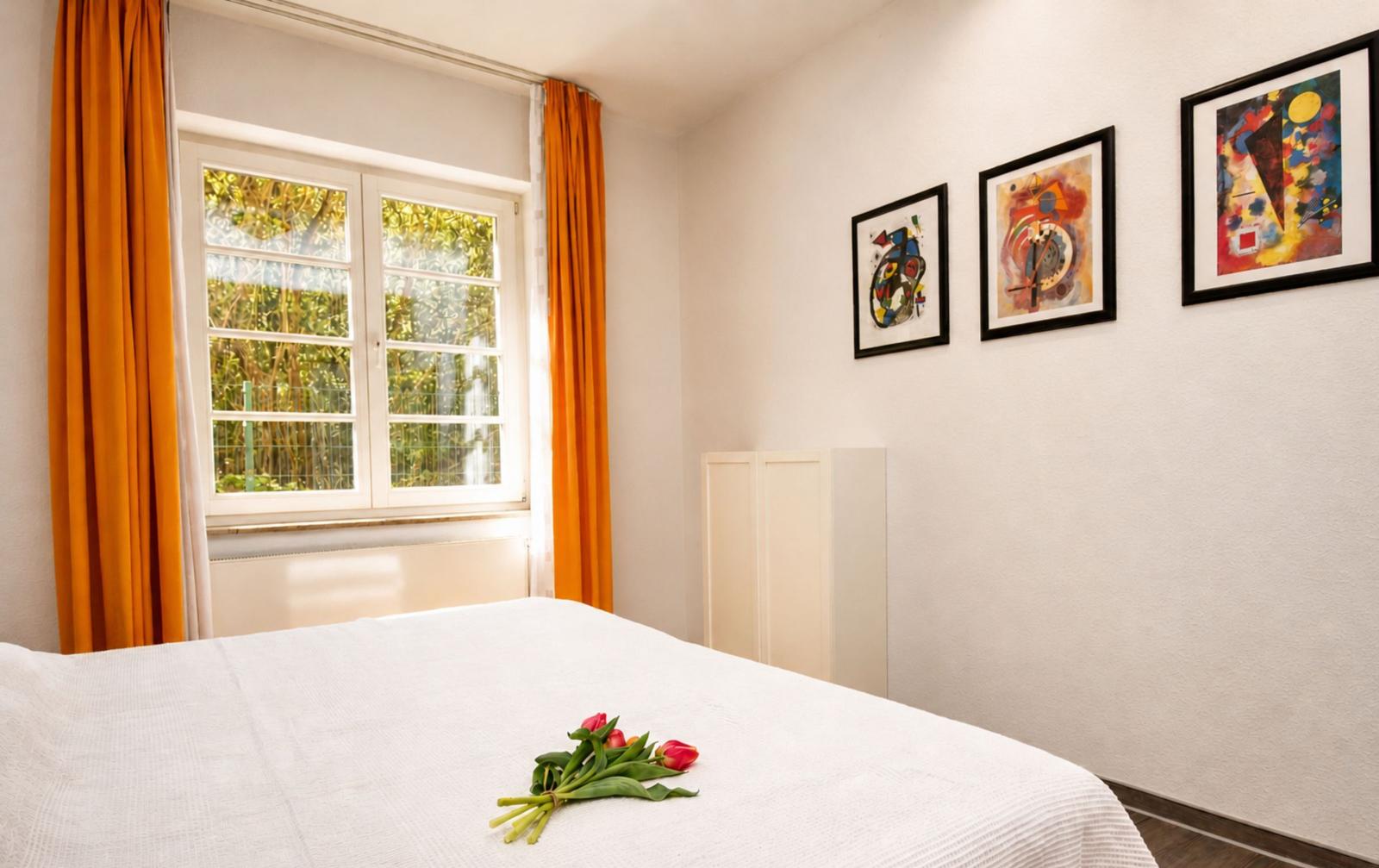 Schlafzimmer mit weißem Bett, orangefarbenen Vorhängen und drei Wandbildern.