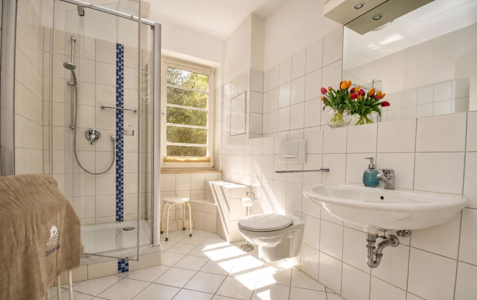 Badezimmer mit Dusche, Toilette und Waschbecken. Fenster, Blumen und Handtuch.