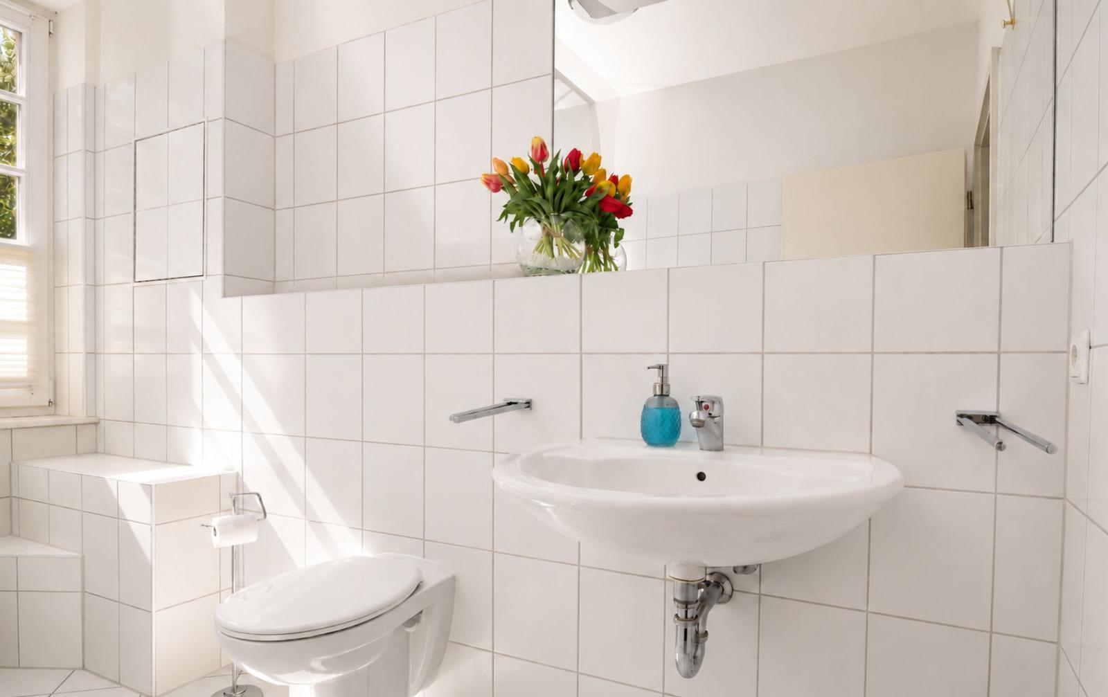 Badezimmer mit Toilette, Waschbecken und Spiegel. Auf dem Waschtisch steht eine Vase mit Tulpen.