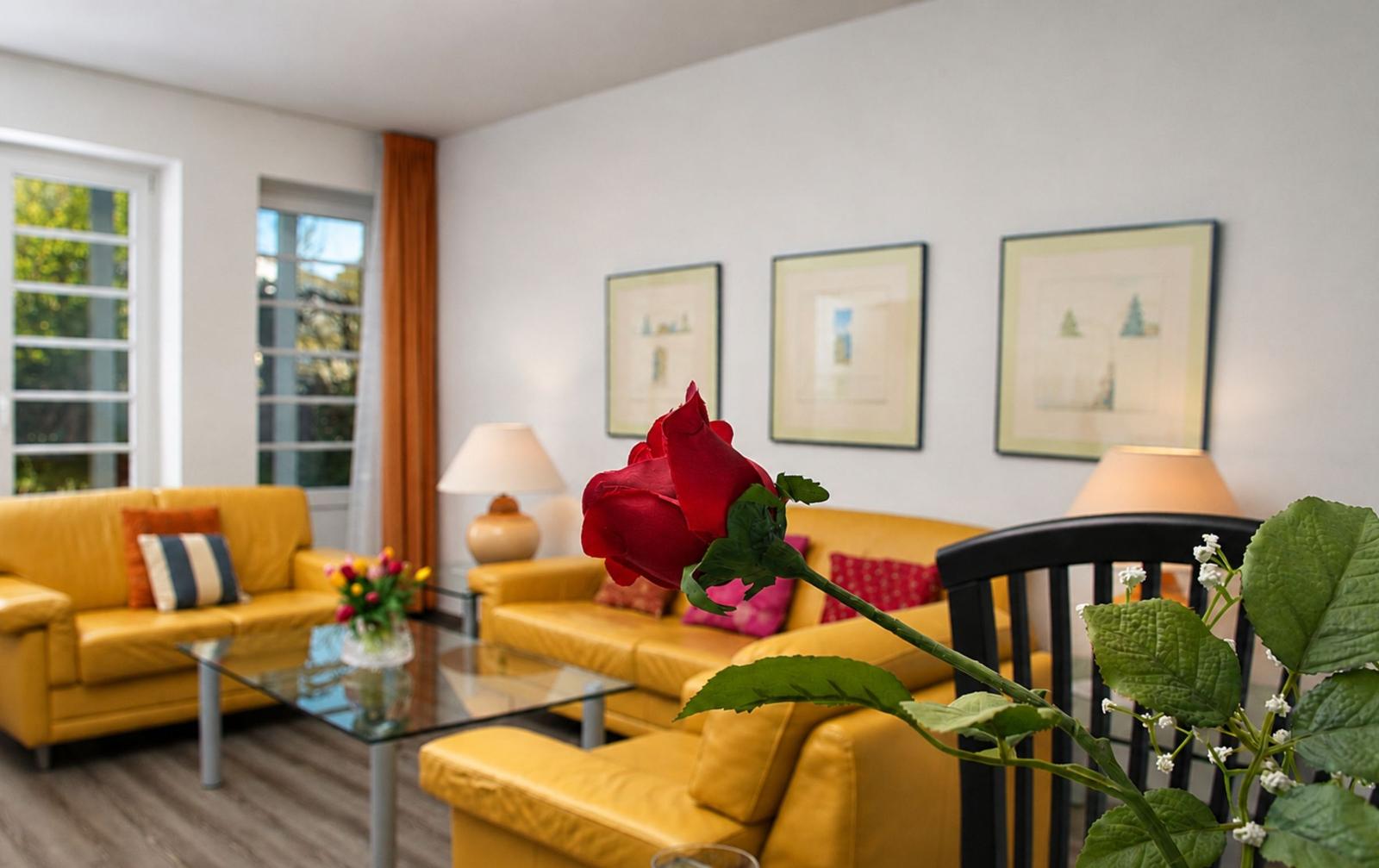 Ein Wohnzimmer mit gelben Sofas, einem Glas-Tisch und einer roten Rose im Vordergrund.