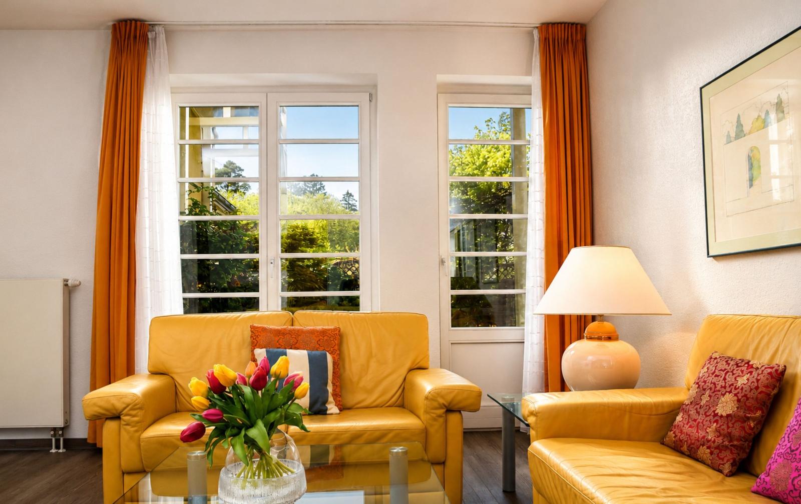 Ein Wohnzimmer mit gelben Ledersofas, einem Glas-Tisch und bunten Kissen. Durch Fenster ist ein Garten sichtbar.