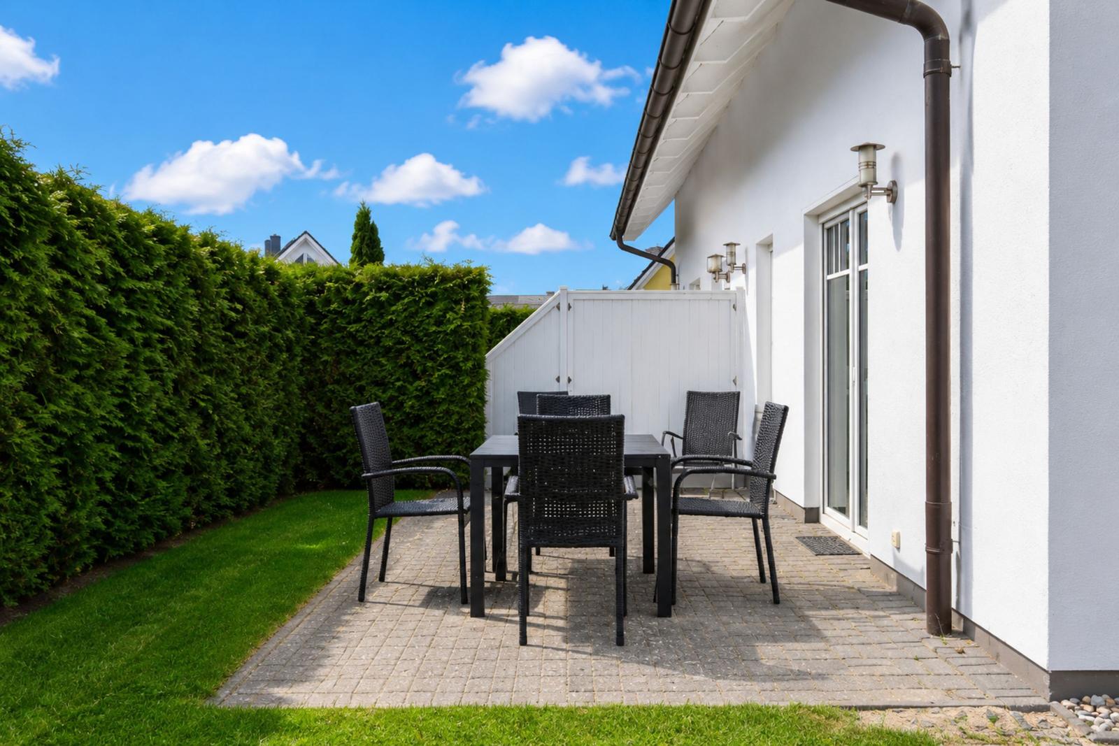 Terrasse mit Tisch und Stühlen neben weißer Hauswand. Hinterhof mit Rasen und Hecke.