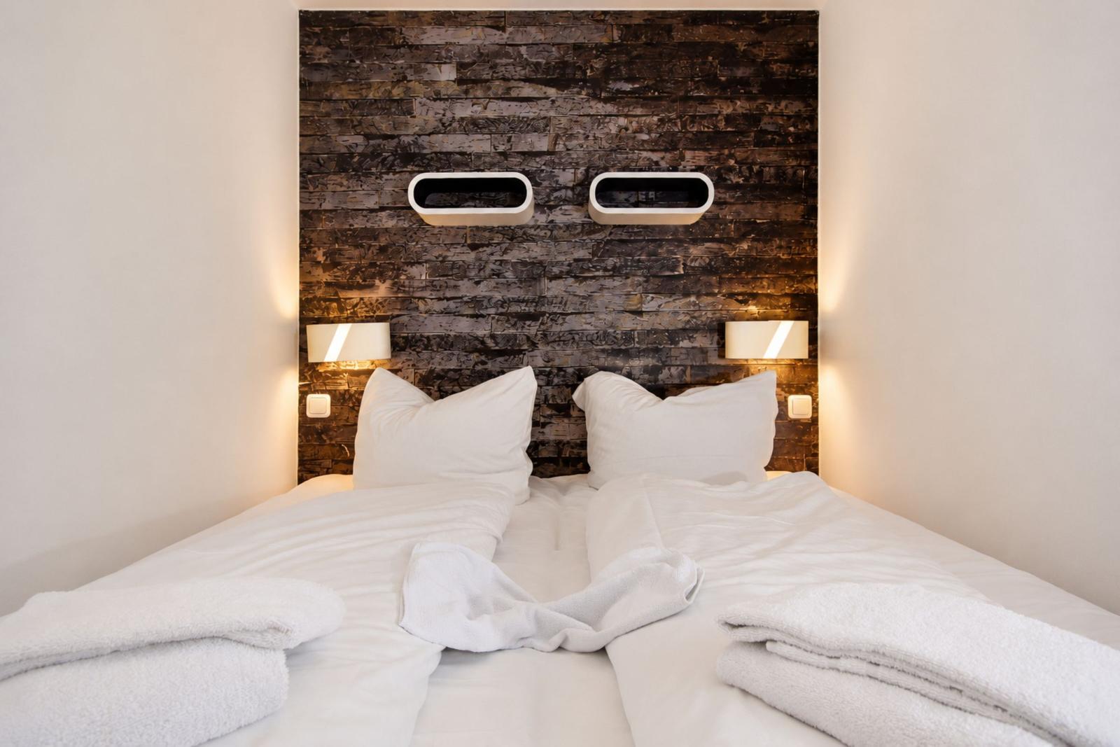 Doppelbett mit weißer Bettwäsche und Handtüchern vor einer Steinwand mit integrierten Lampen.