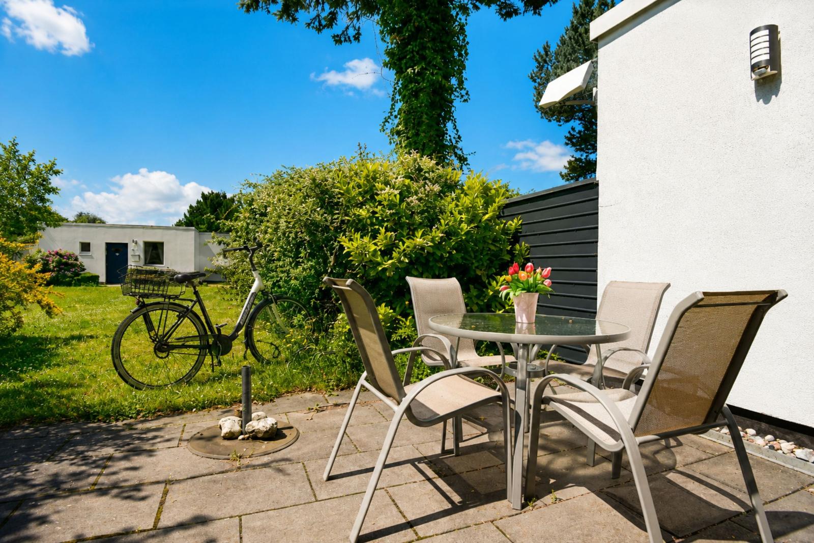 Terrasse mit Tisch, Stühlen und Fahrrad. Hinterhof mit Grünflächen und Hecke.