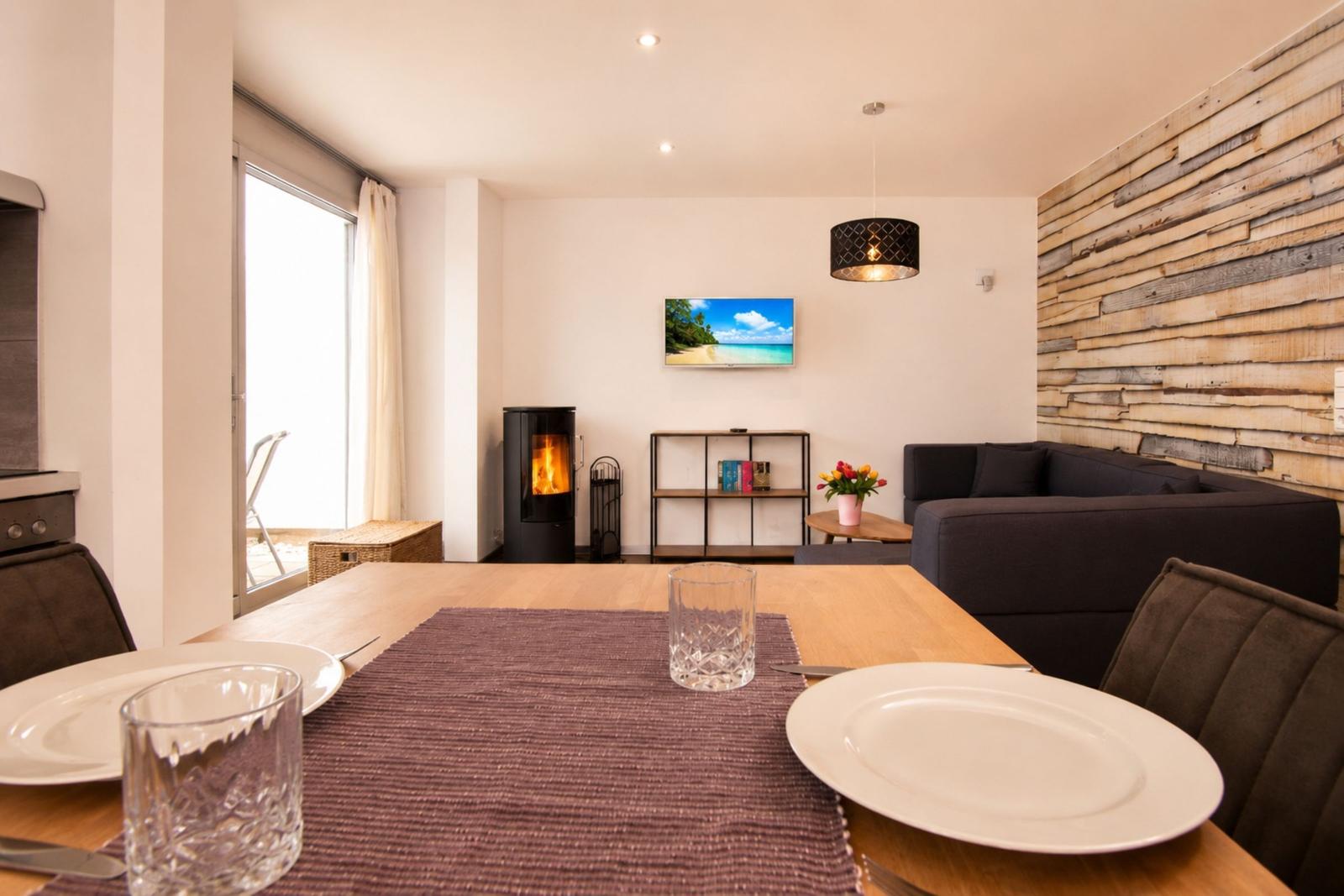 Esszimmer mit Tisch, Stühlen und Fernseher. Im Hintergrund steht ein Sofa und ein Kaminofen.