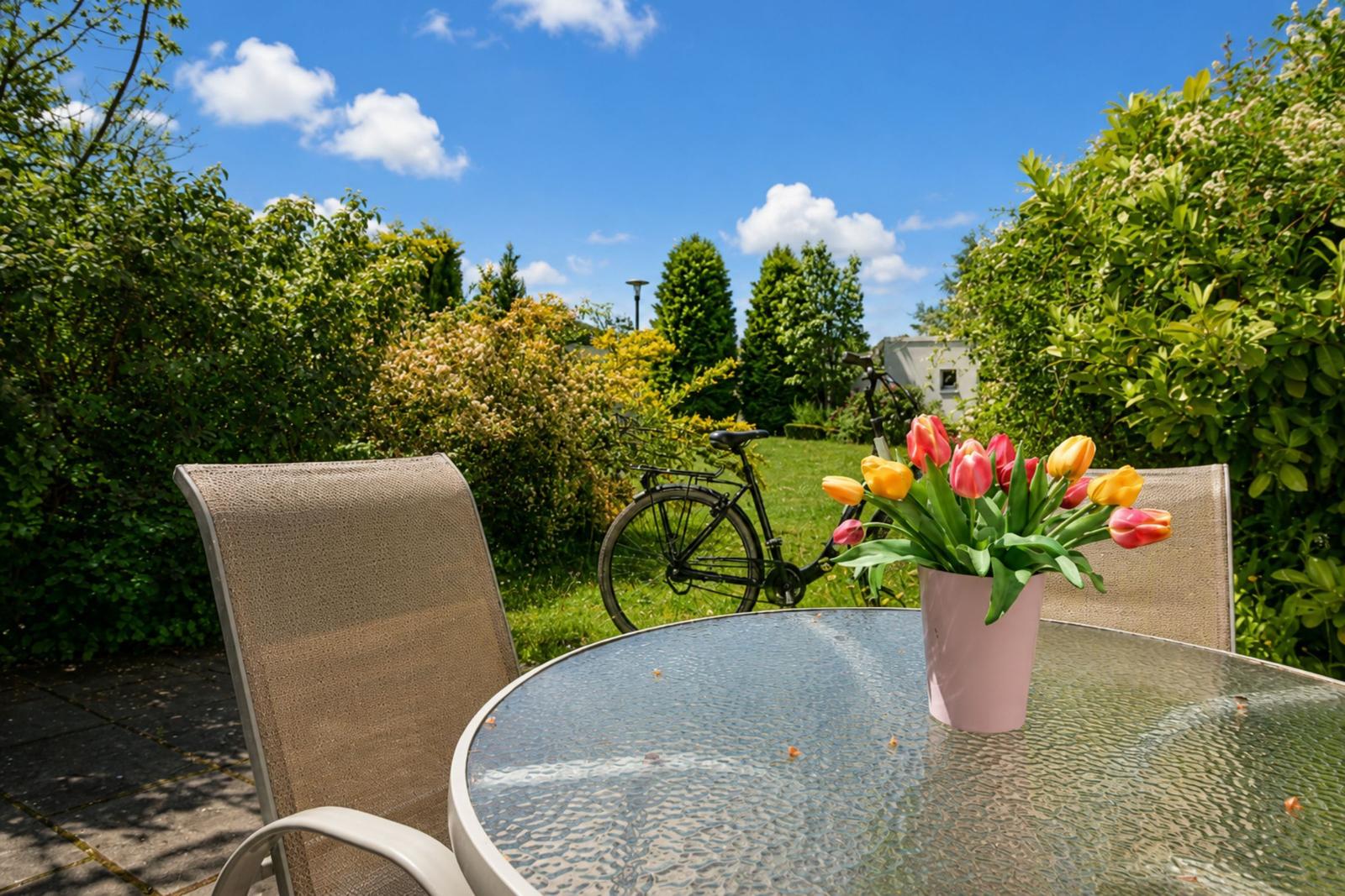Runder Glastisch mit Stühlen und Tulpen im Garten. Fahrrad und grüne Hecke im Hintergrund.