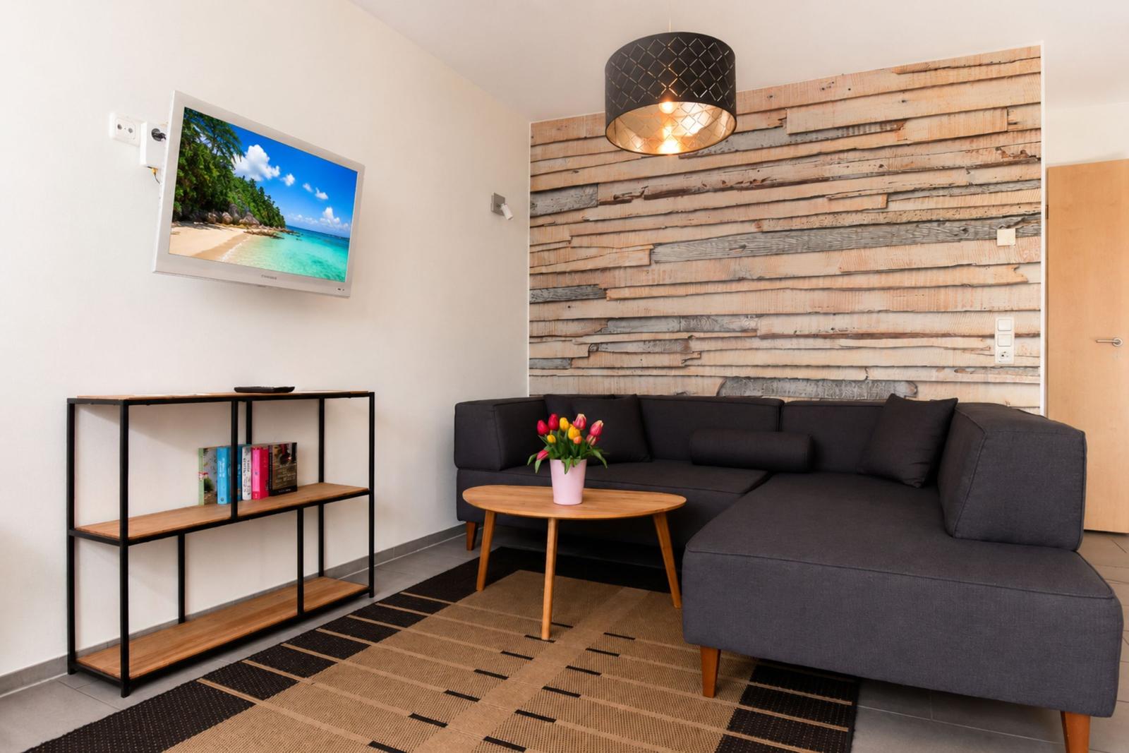 Wohnzimmer mit Sofa, Fernseher und Holzwand. Tisch mit Blumen und Regal mit Büchern.