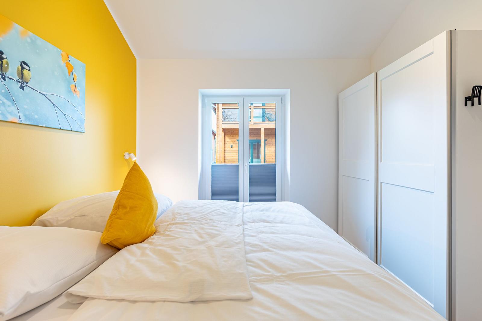 Schlafzimmer mit Bett, gelber Wand und Bild. Tür führt nach draußen.