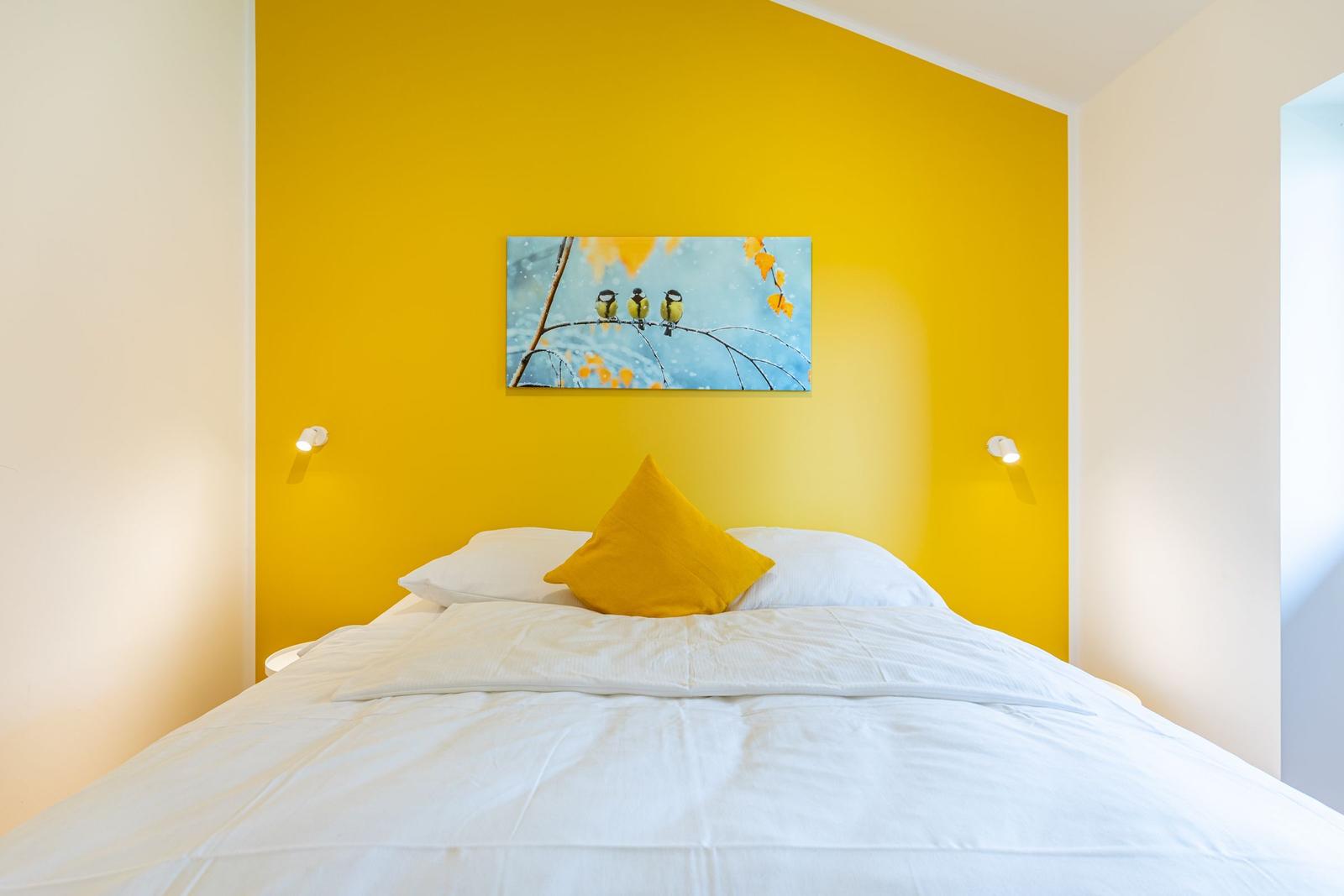 Schlafzimmer mit gelber Wand, Bett mit weißer Bettwäsche und gelbem Kissen, Wandbild mit Vögeln.