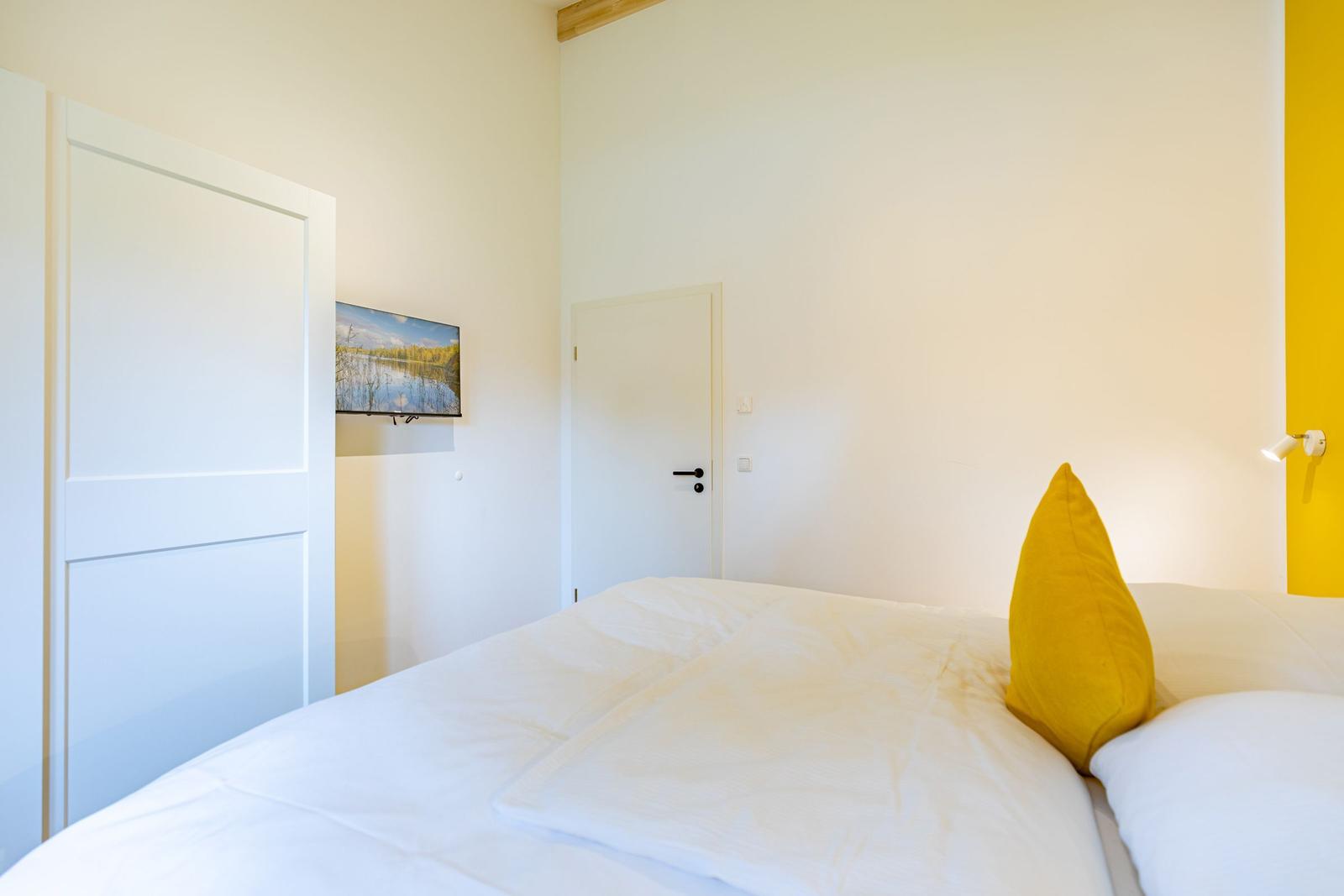 Schlafzimmer mit Bett, gelber Kissen und Fernseher an der Wand.