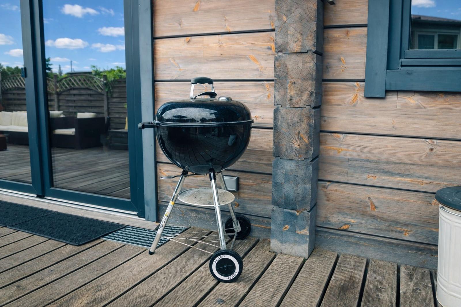 Schwarzer Grill auf Holzterrasse neben Holzwand und Glastür.